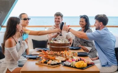 Cinco amigos brindan en la cubierta alrededor de una mesa con langosta, sushi, ostras y fruta, con el mar detrás