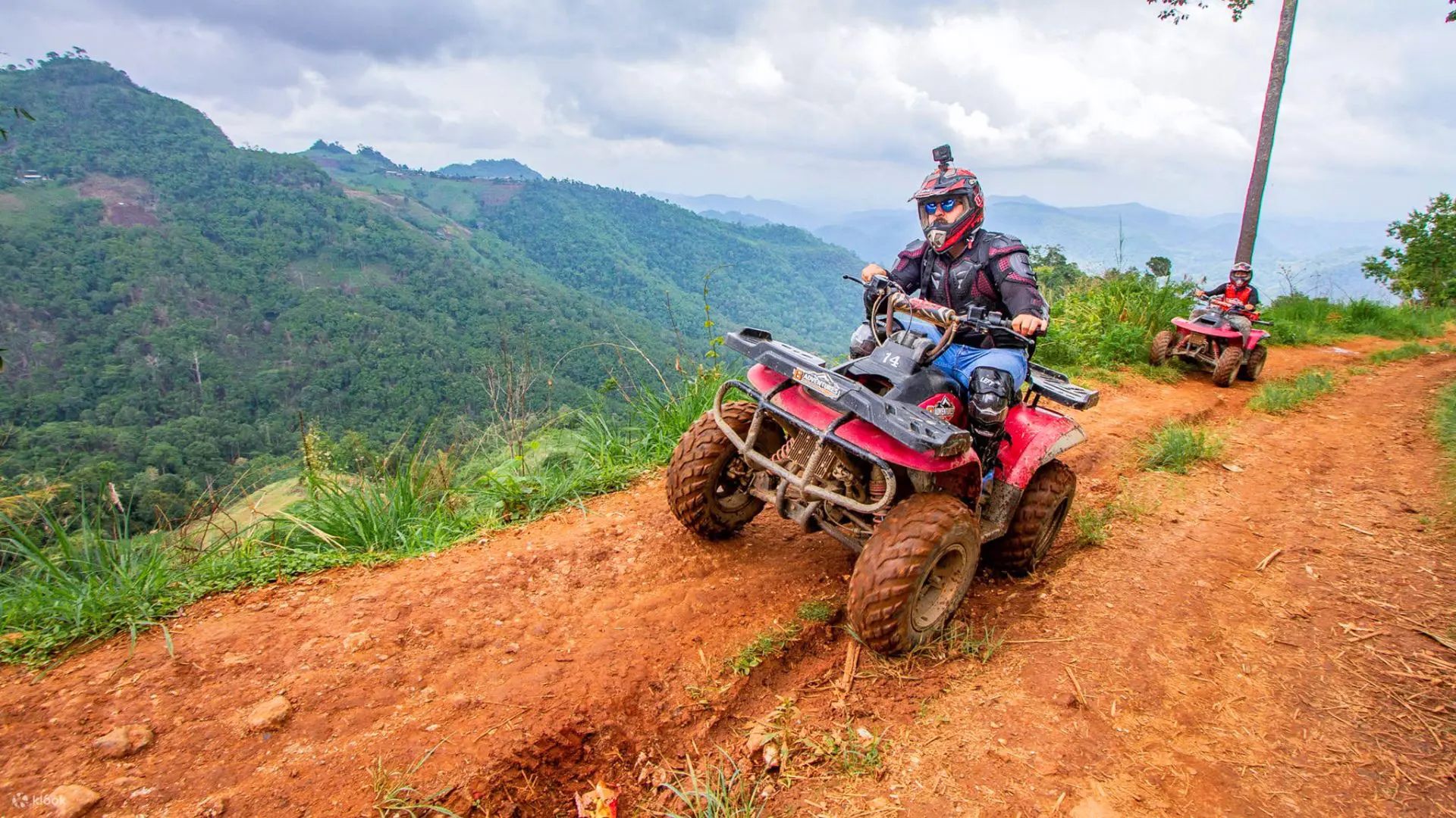 Red quad climbing a red dirt track in the mountains, with another rider behind and valleys in the background