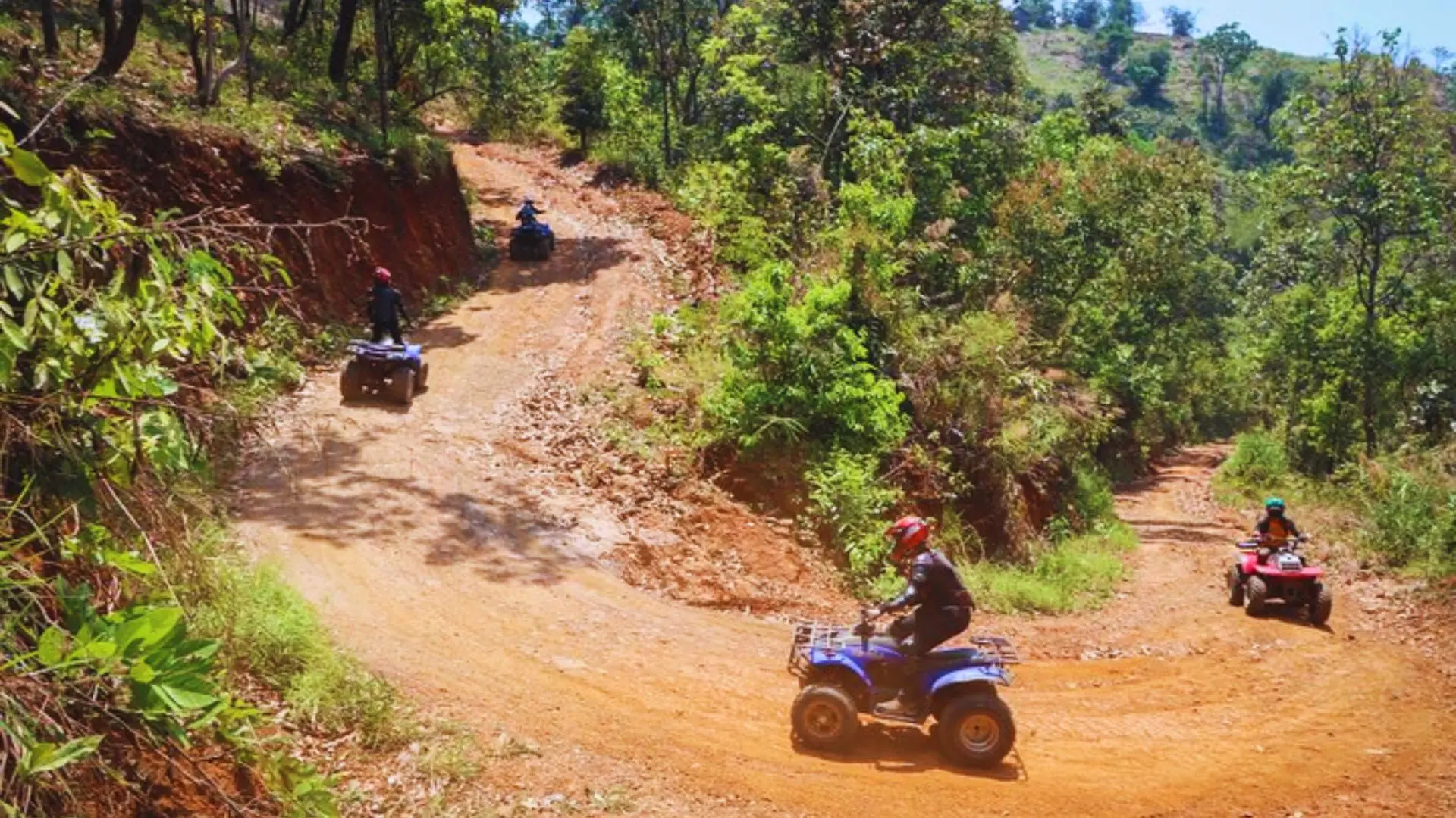 Four quads ride an orange dirt track winding in curves along a green forested slope