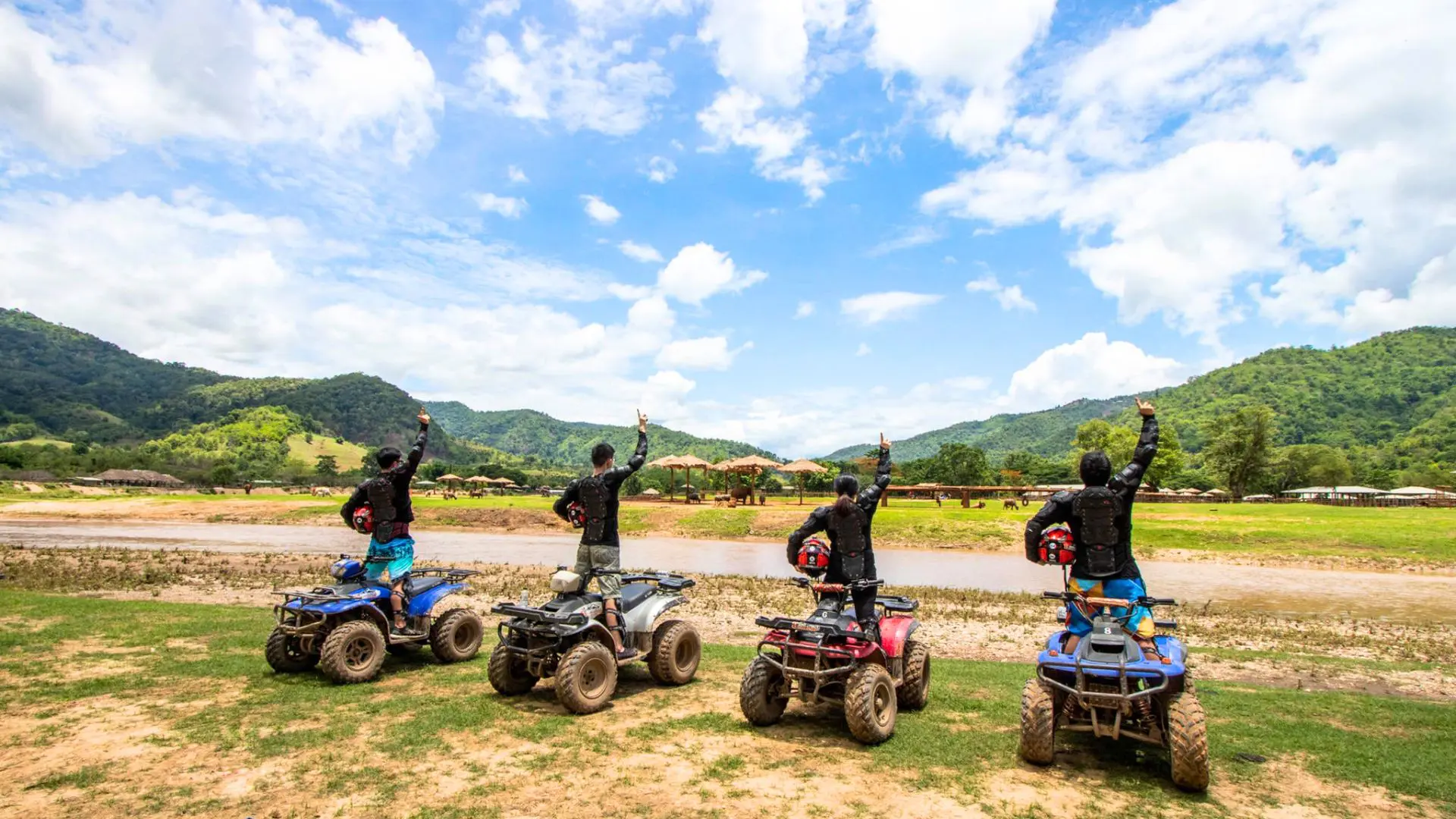 Four riders standing on their quads raise an arm before a river and a green valley ringed by mountains