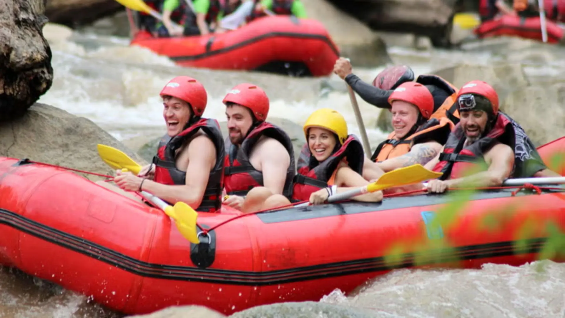 Six people in helmets laugh in a red rafting raft, one holding a paddle aloft, with other rafts in the background