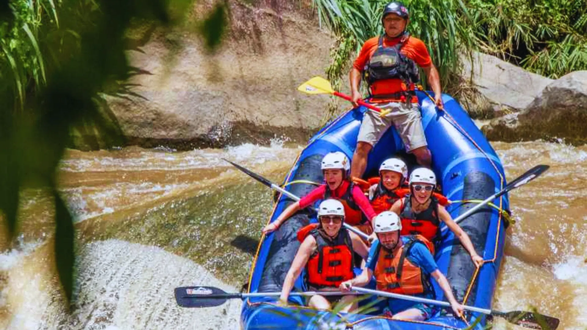 Blue raft clearing a drop in the water while the guide, standing at the stern, directs five helmeted paddlers