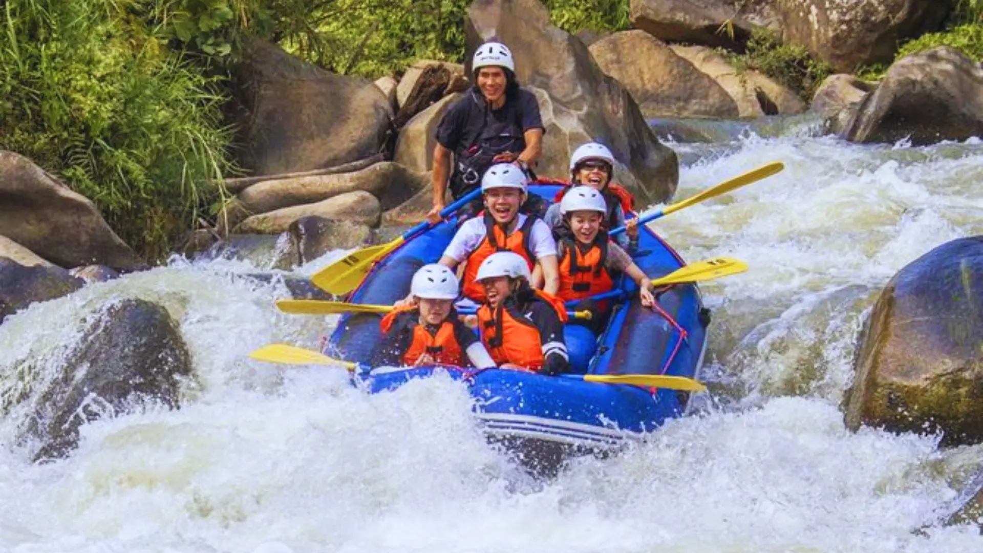 Five paddlers in white helmets shout in a blue raft amid rapids and big rocks, with the guide in black standing behind