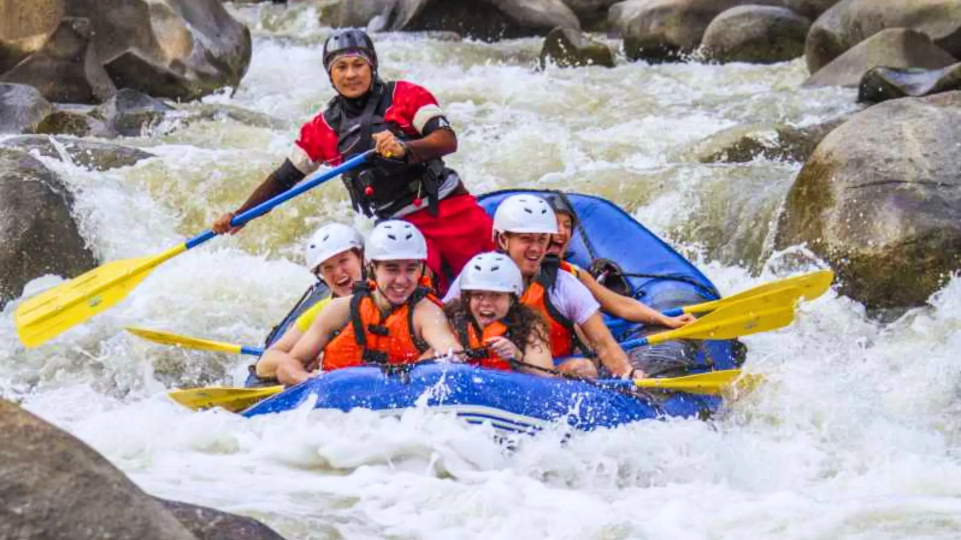 Guide in a red T-shirt paddles at the stern as the group in helmets and life jackets runs rapids between rocks