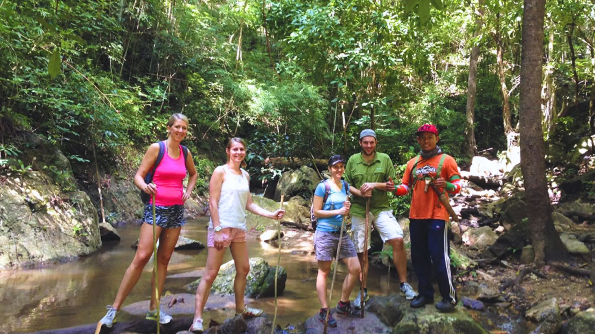 Four travelers and a local guide pose with bamboo sticks on the rocks of a jungle stream