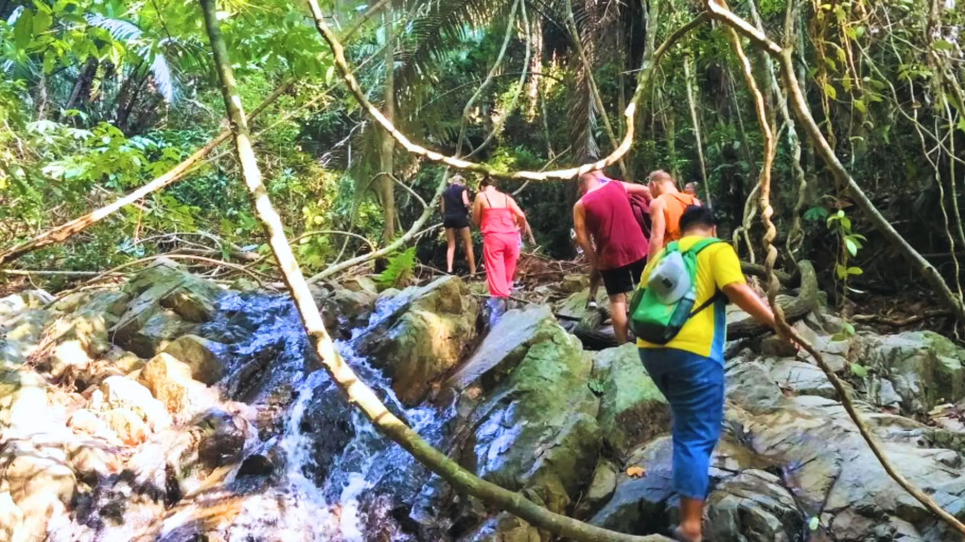 Hikers climb the rocks of a jungle stream with a waterfall; a man with a green backpack brings up the rear