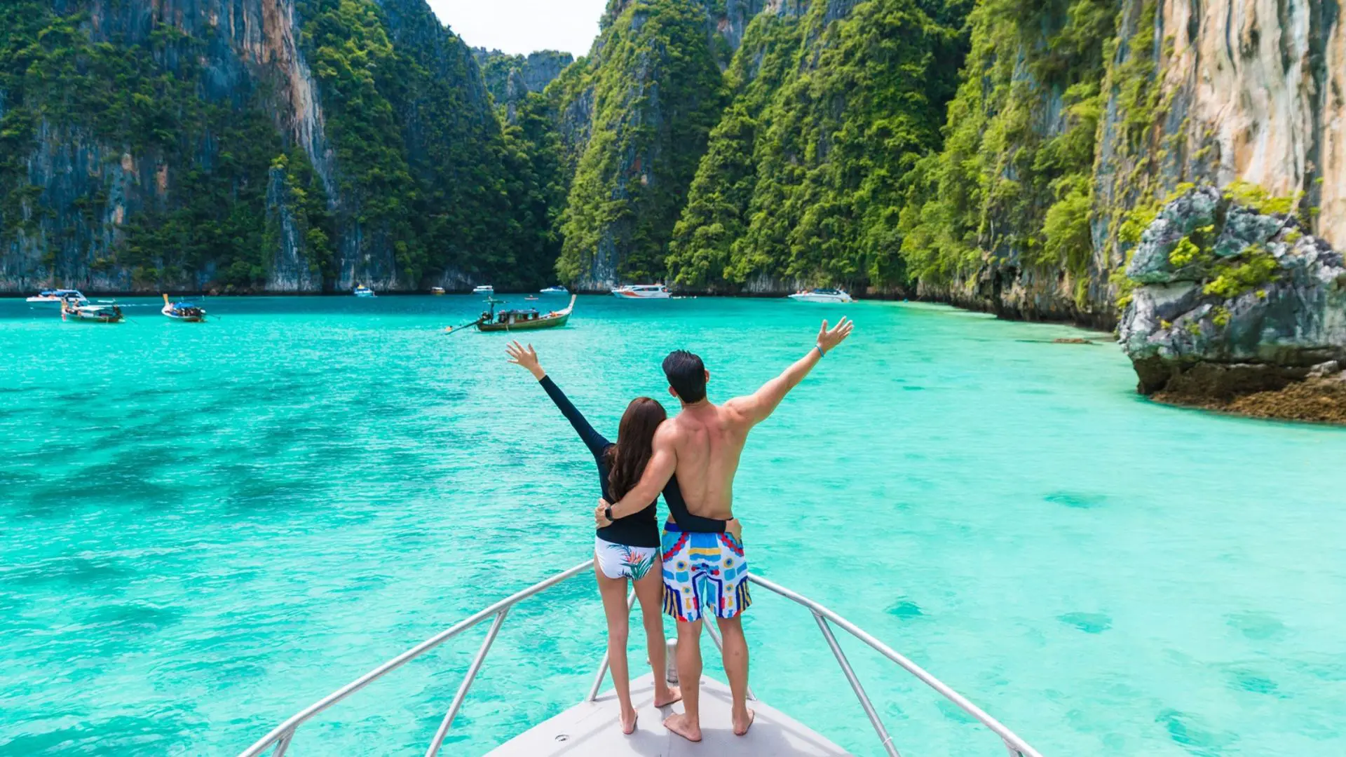Casal abraçado na proa de uma lancha levanta os braços diante de uma lagoa turquesa com barcos ao fundo