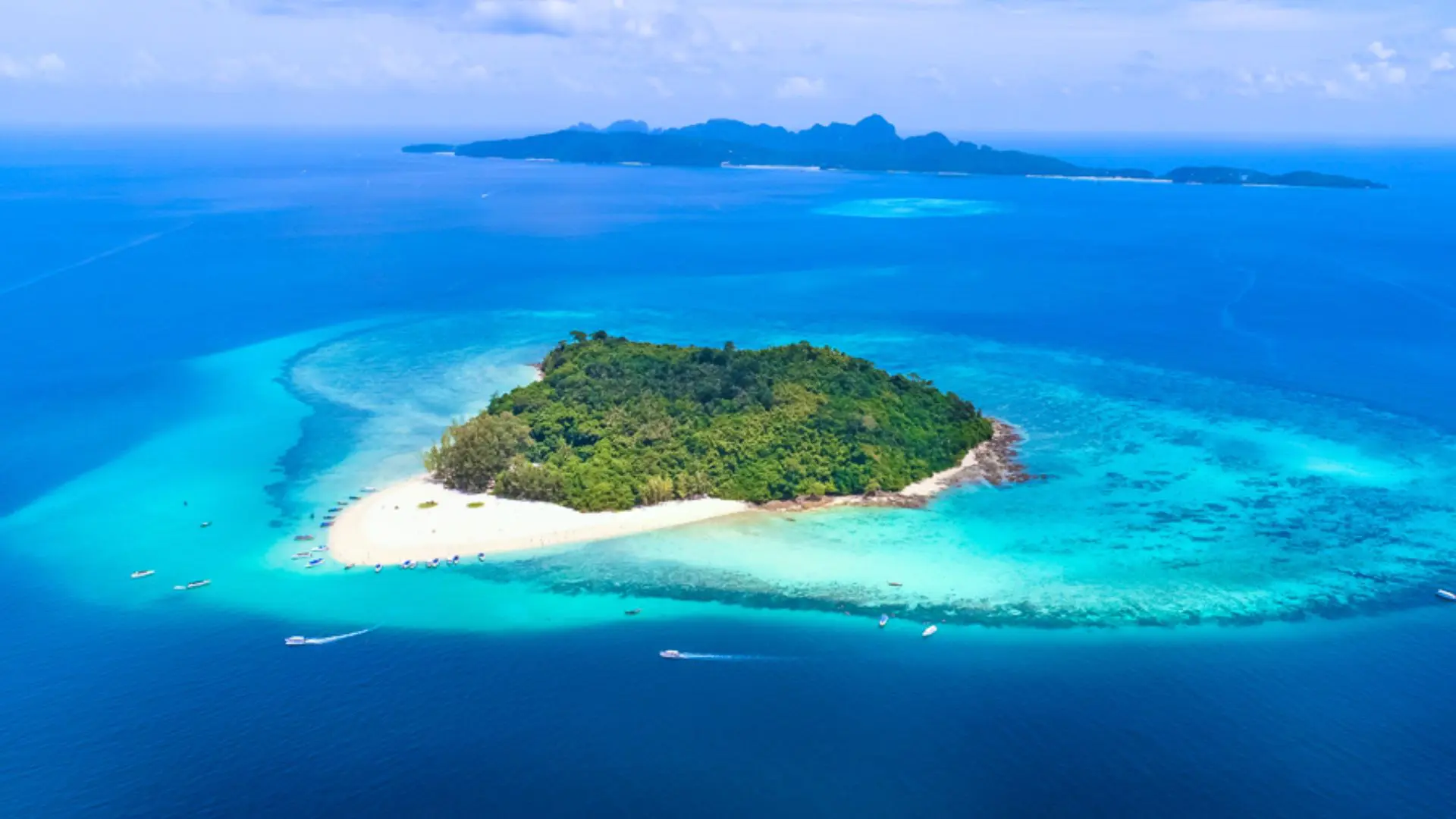 Vue aérienne d’une petite île boisée entourée d’un récif turquoise, avec une île plus grande au fond