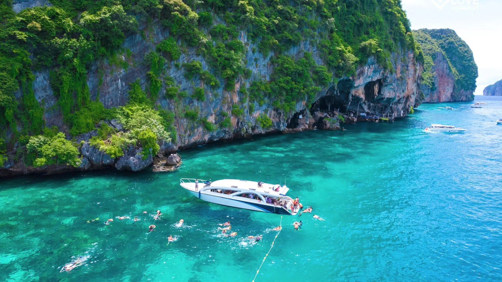 Grupo numeroso faz snorkel junto a uma lancha branca fundeada ao pé de uma falésia com selva, visto do ar