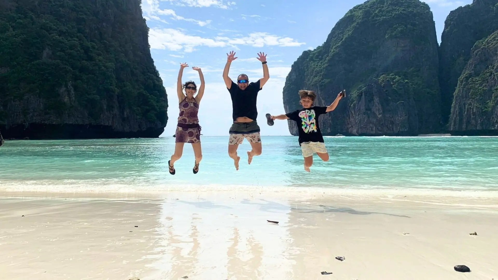 Three travelers jump at the same time on the wet sand of Maya Bay, with the cliffs behind them