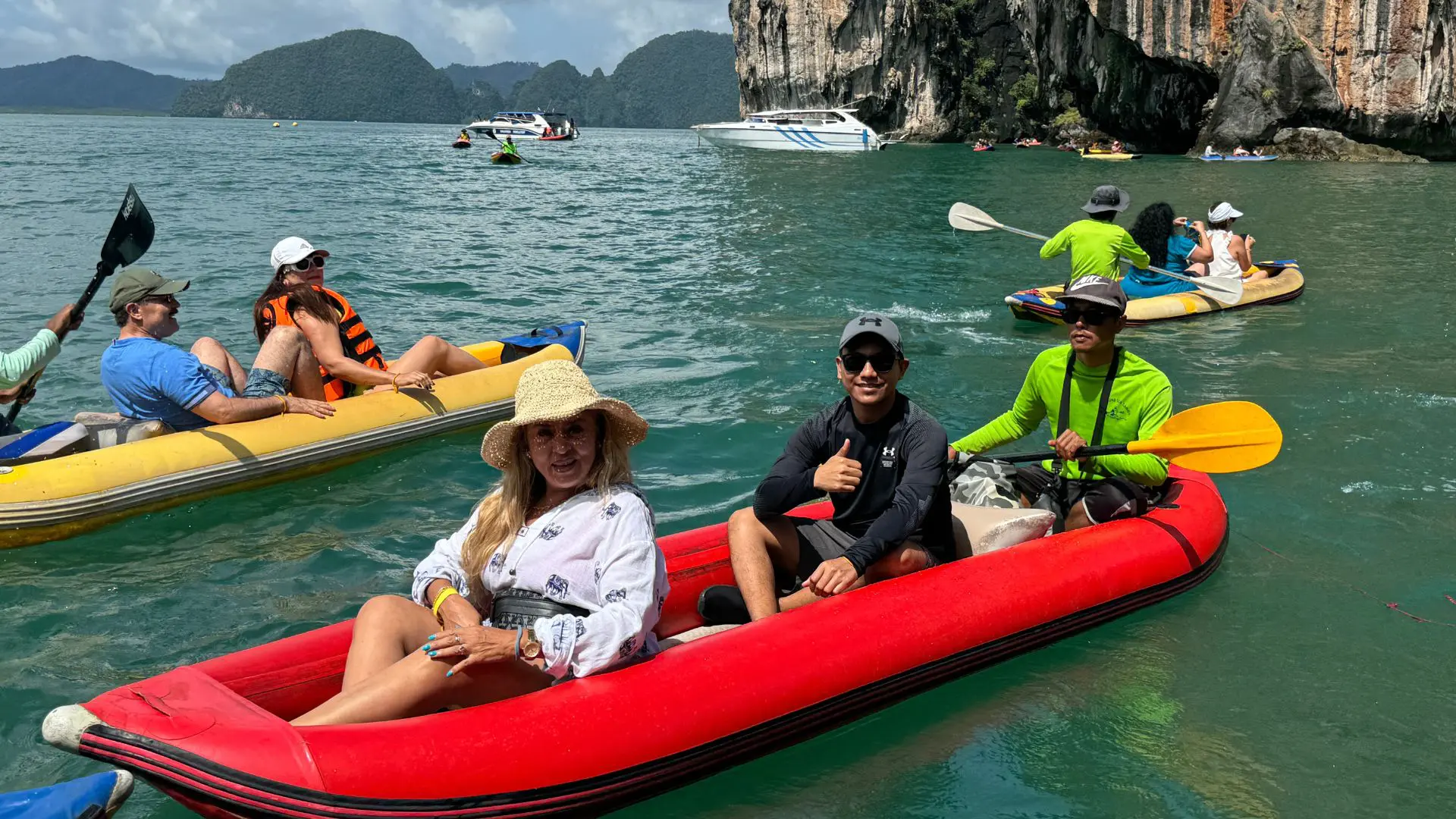 Mujer con sombrero de paja y joven con el pulgar arriba en una canoa roja; detrás, otra canoa amarilla y lanchas