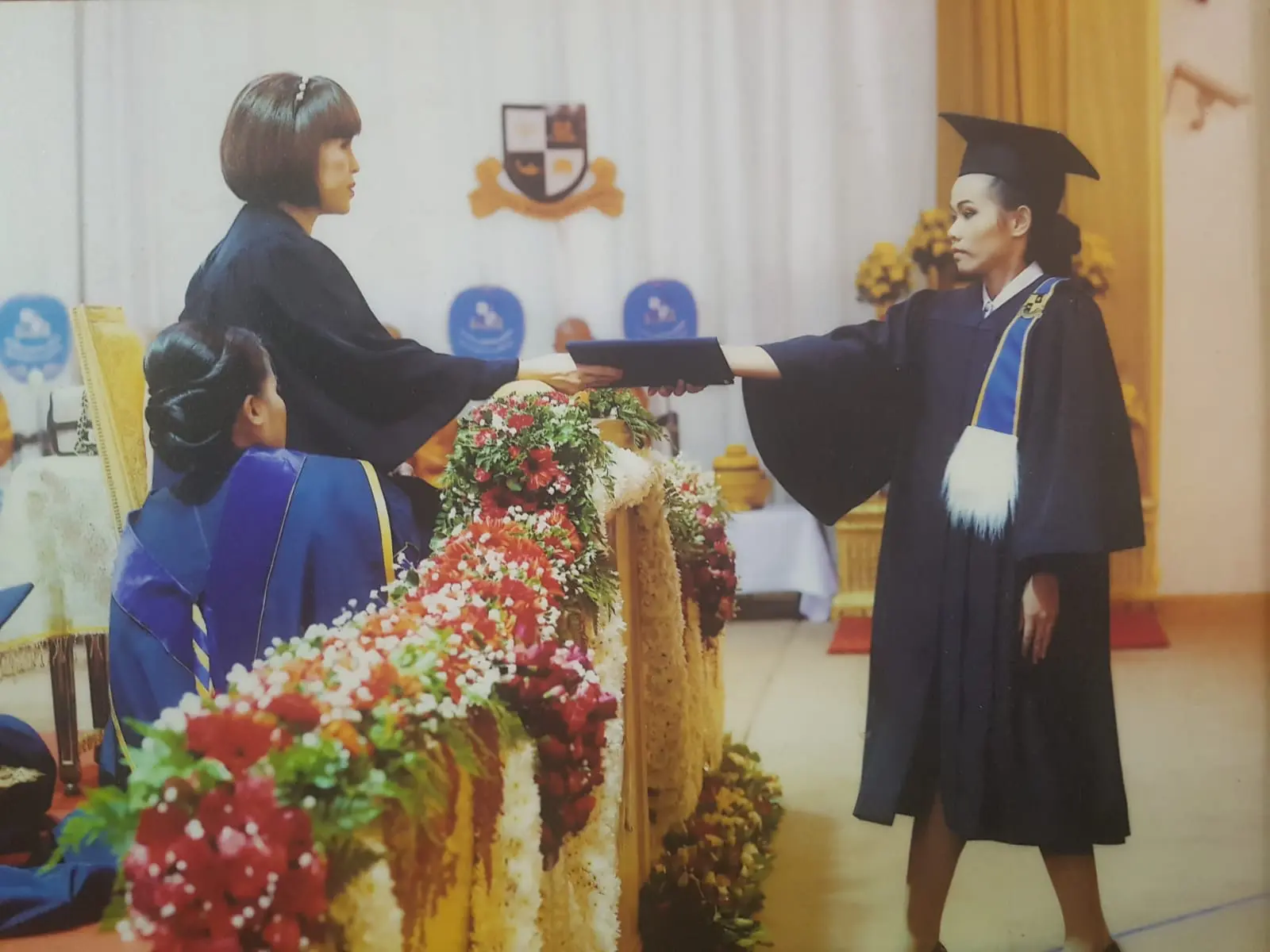 Graduación: una mujer con toga y birrete recibe su diploma en un estrado adornado con flores rojas y blancas