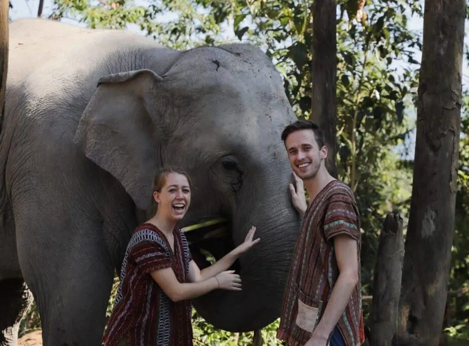 Um casal com túnicas tailandesas ri junto a um elefante enquanto ele acaricia a tromba do animal, entre árvores