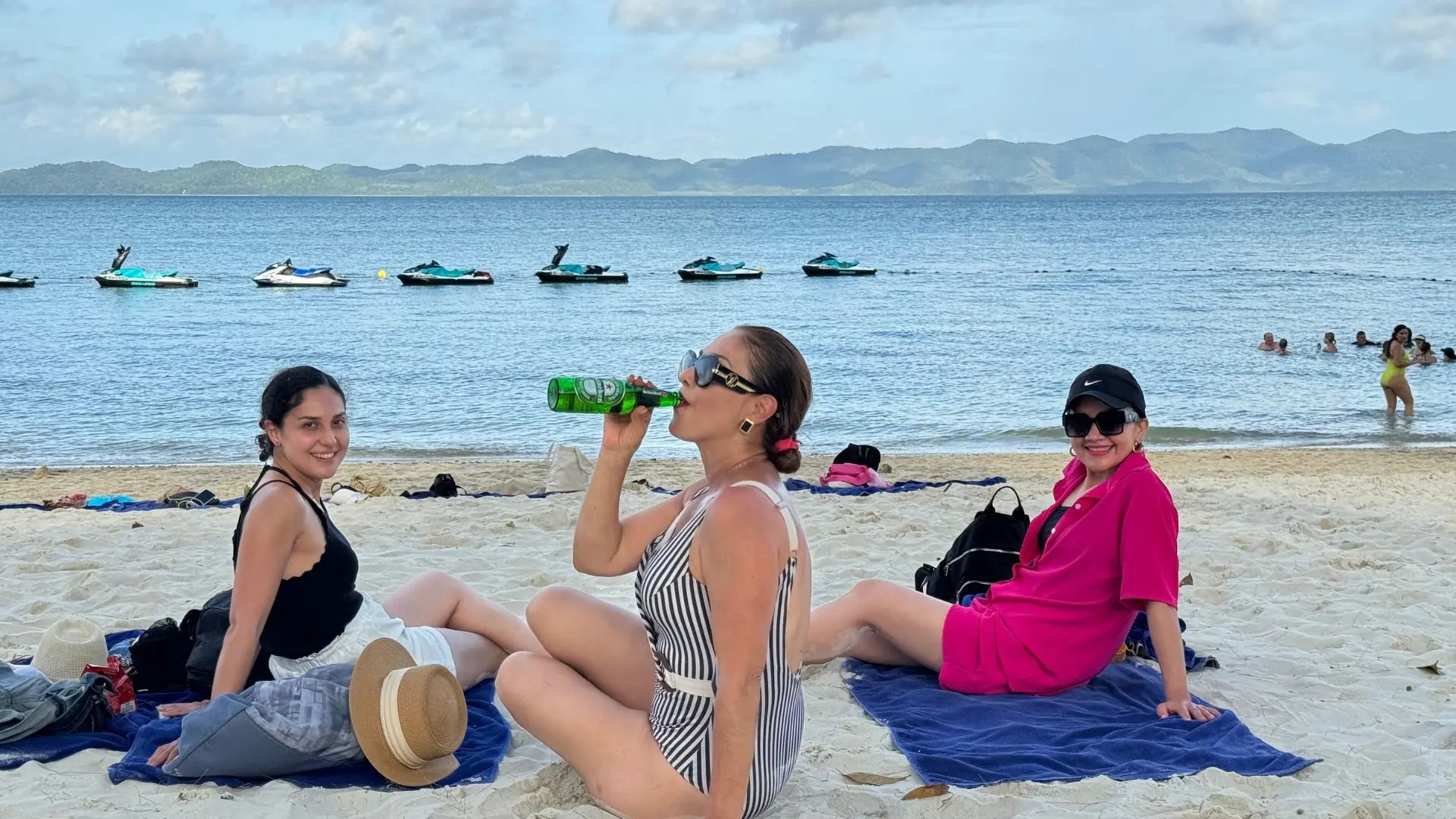 Tres mujeres descansan en la arena frente al mar, con motos de agua fondeadas y montañas verdes al fondo