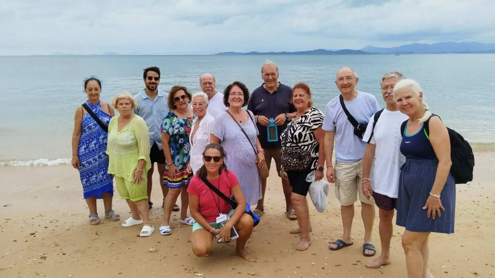 Grupo de trece viajeros posando en una playa de arena, con el mar y montañas al fondo en un día nublado