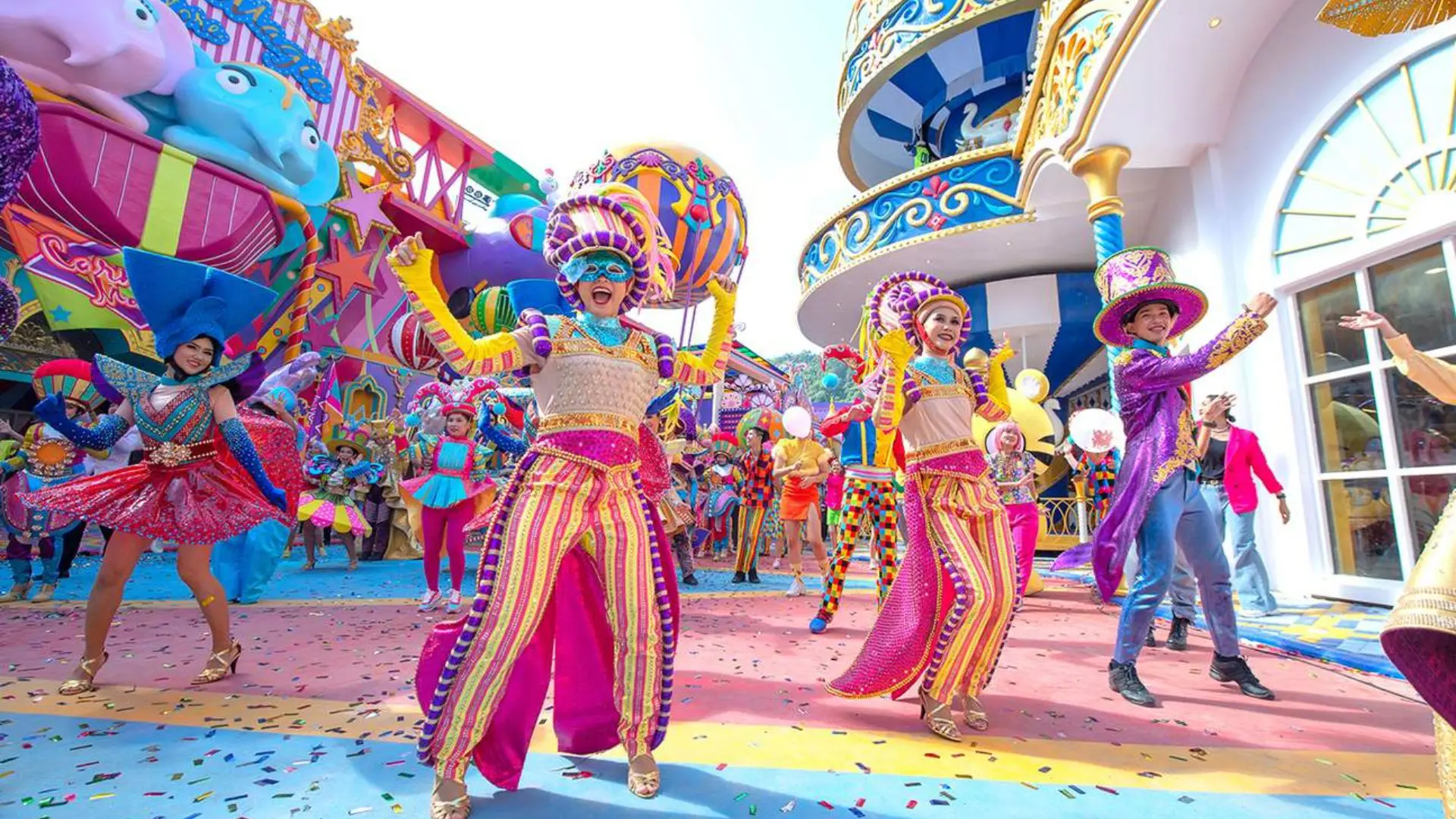 Masked dancer in striped pants leads the parade amid confetti, with more dancers and an elephant float
