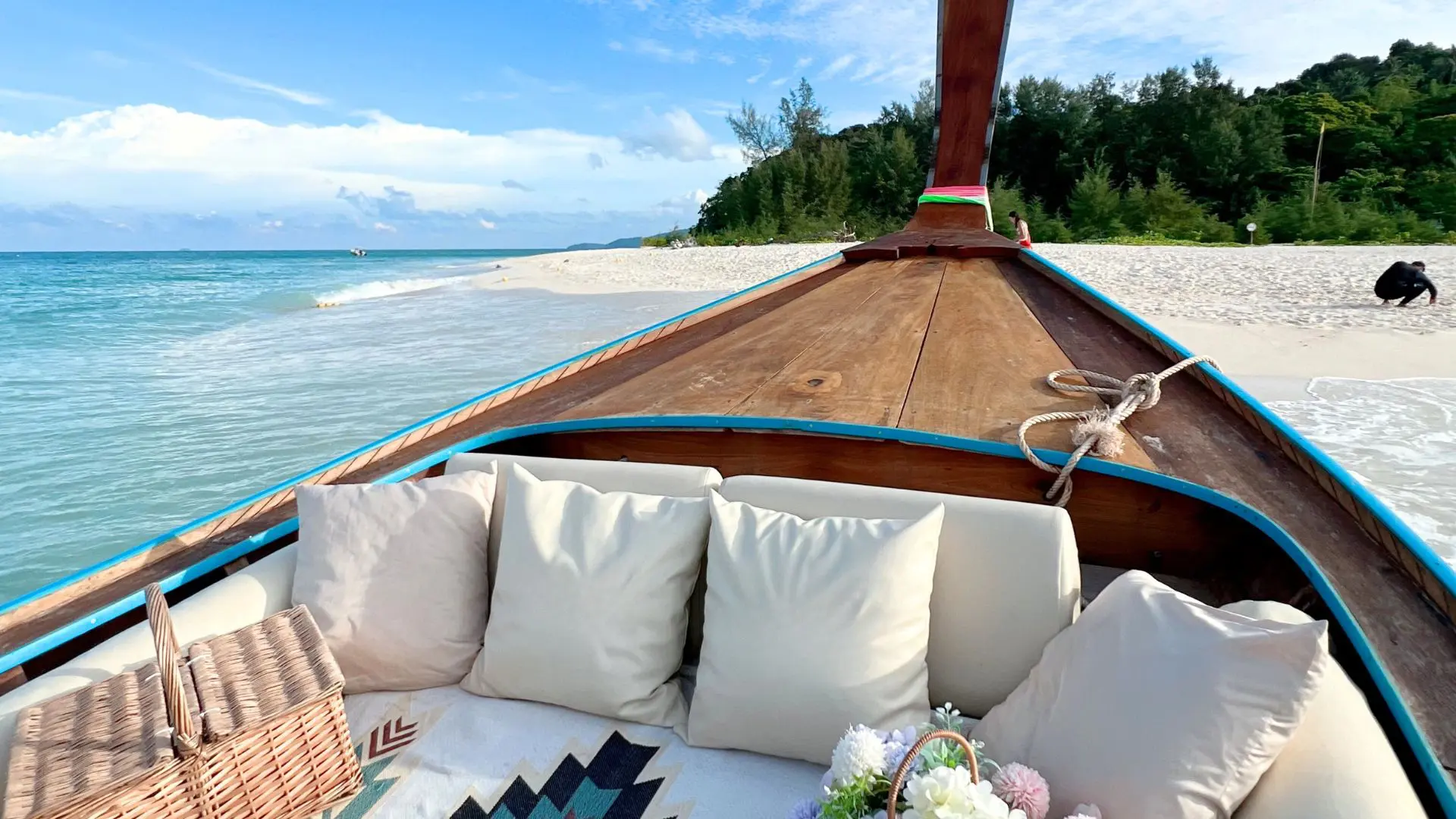 Interior de um barco de cauda longa com almofadas brancas, cesto de piquenique e flores, varado numa praia de areia clara