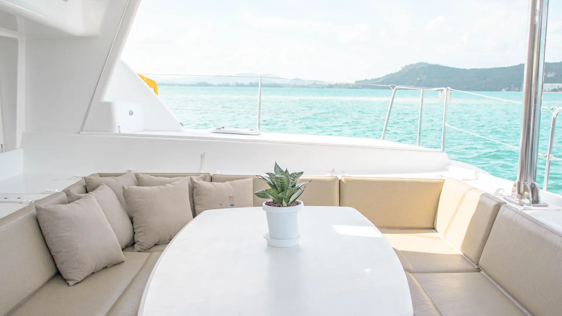 Outdoor lounge of a catamaran with beige sofas and a white table with a plant, facing the turquoise sea