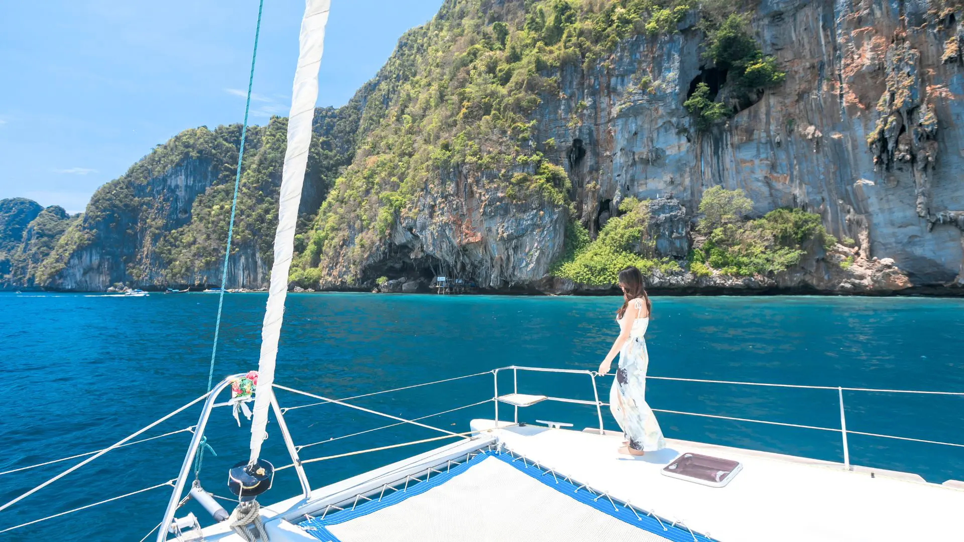 Woman in a long dress walking along the bow of a catamaran in front of a huge limestone cliff