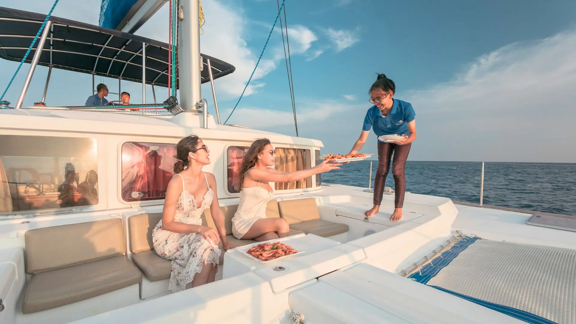 A crew member serves a tray of shrimp to two female passengers on the catamaran deck in sunset light