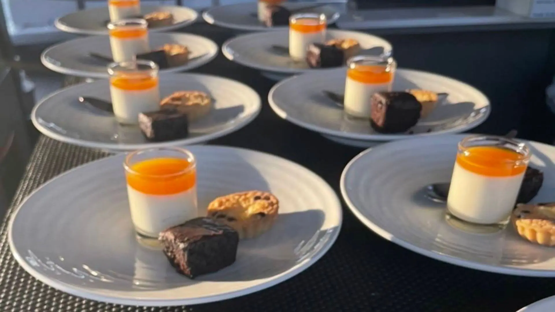 Platos de postre en fila: panna cotta con cobertura naranja, brownie y tartaleta, con luz del atardecer