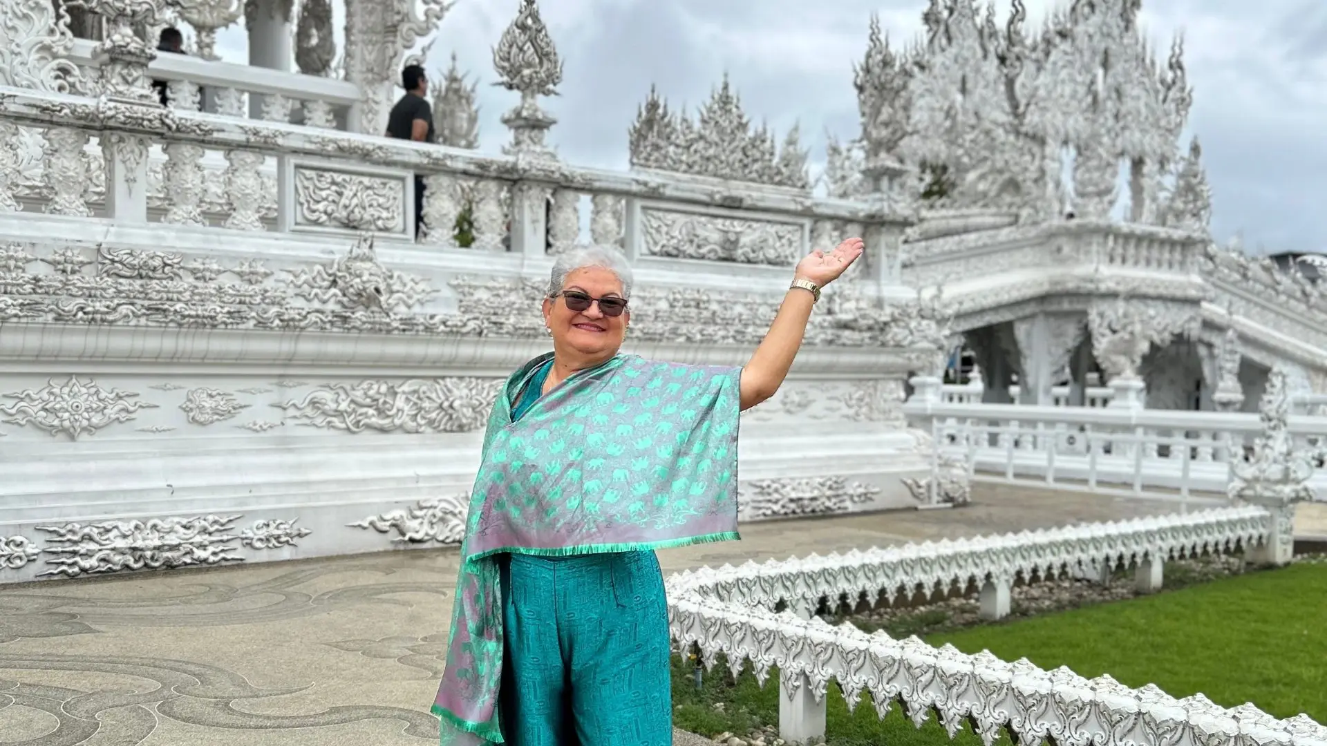 A white-haired traveler in a green shawl raises her arm before the carved balustrade of the White Temple