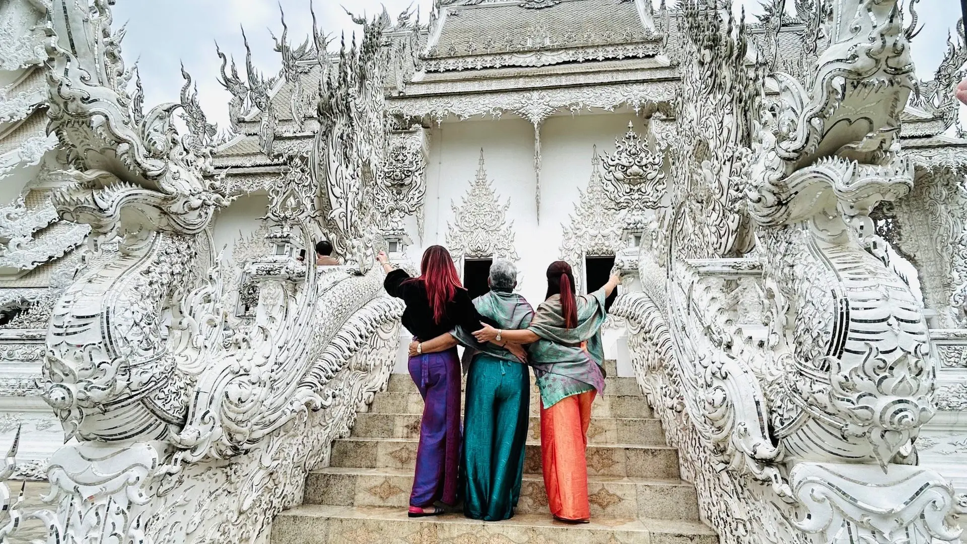 Three female travelers hugging, seen from behind, on the White Temple stairway between white plaster nagas