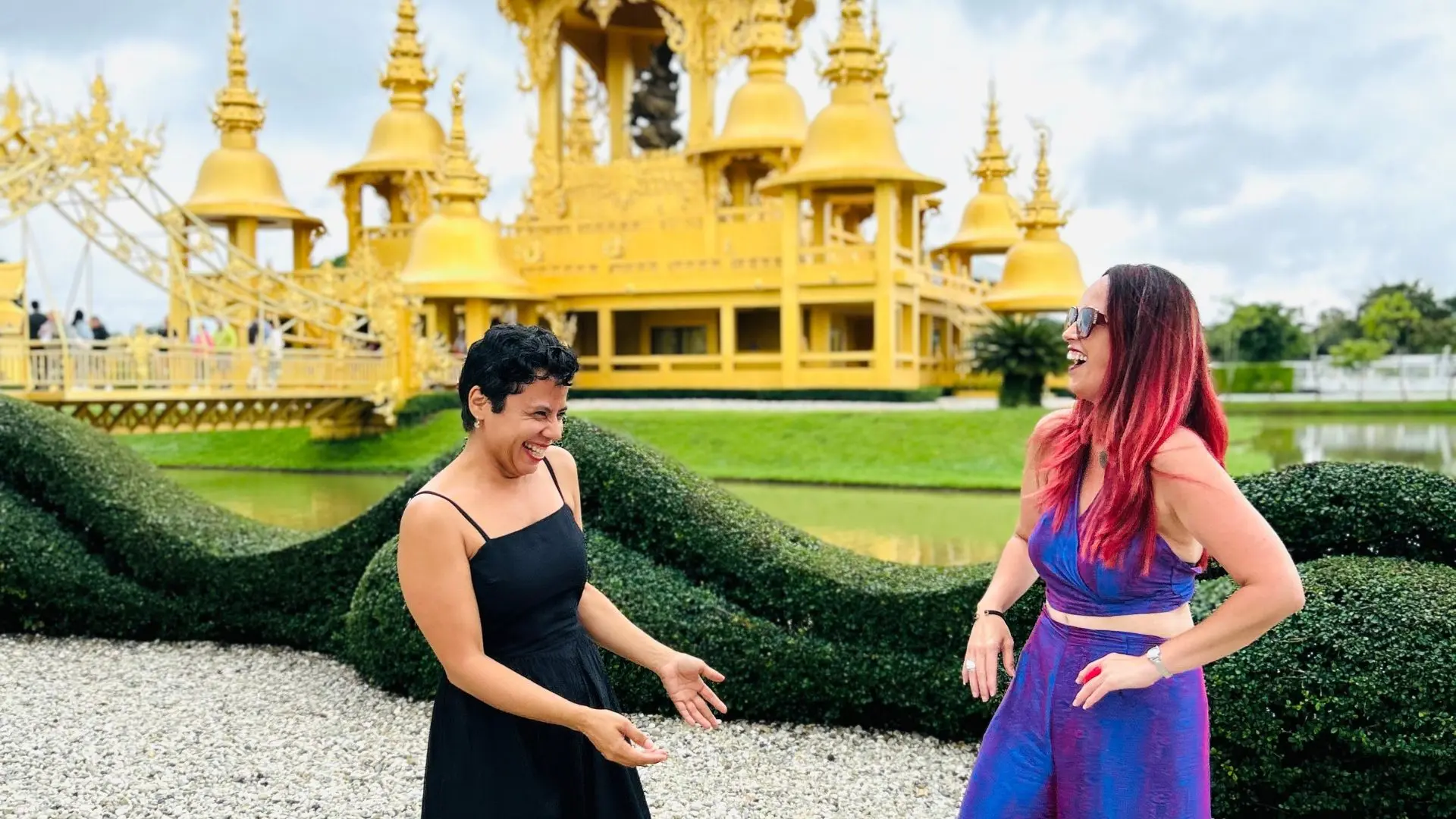 Two female travelers laugh in front of the golden pavilion of Wat Rong Khun, with clipped hedges and a cloudy sky