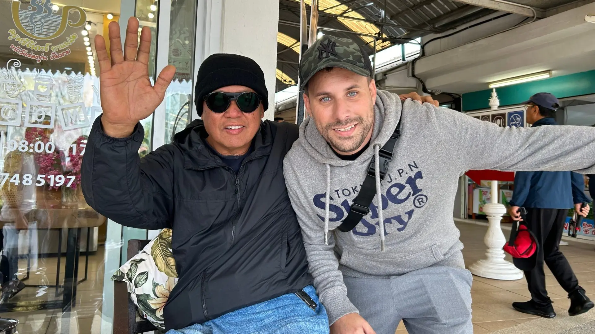 A local man in a cap and sunglasses waves at the camera, arm around a traveler, in front of a shop