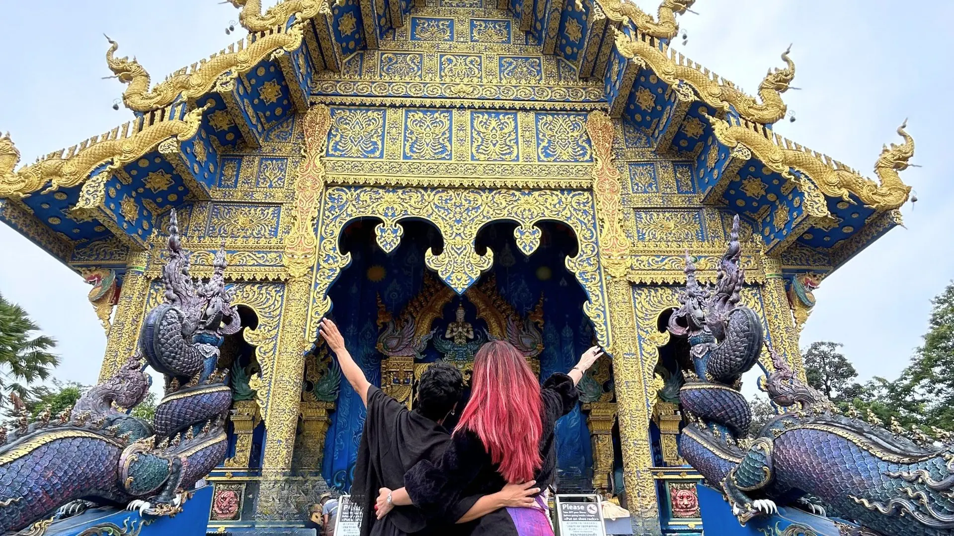Two friends arm in arm, seen from behind, raise an arm before the blue and gold facade of Wat Rong Suea Ten