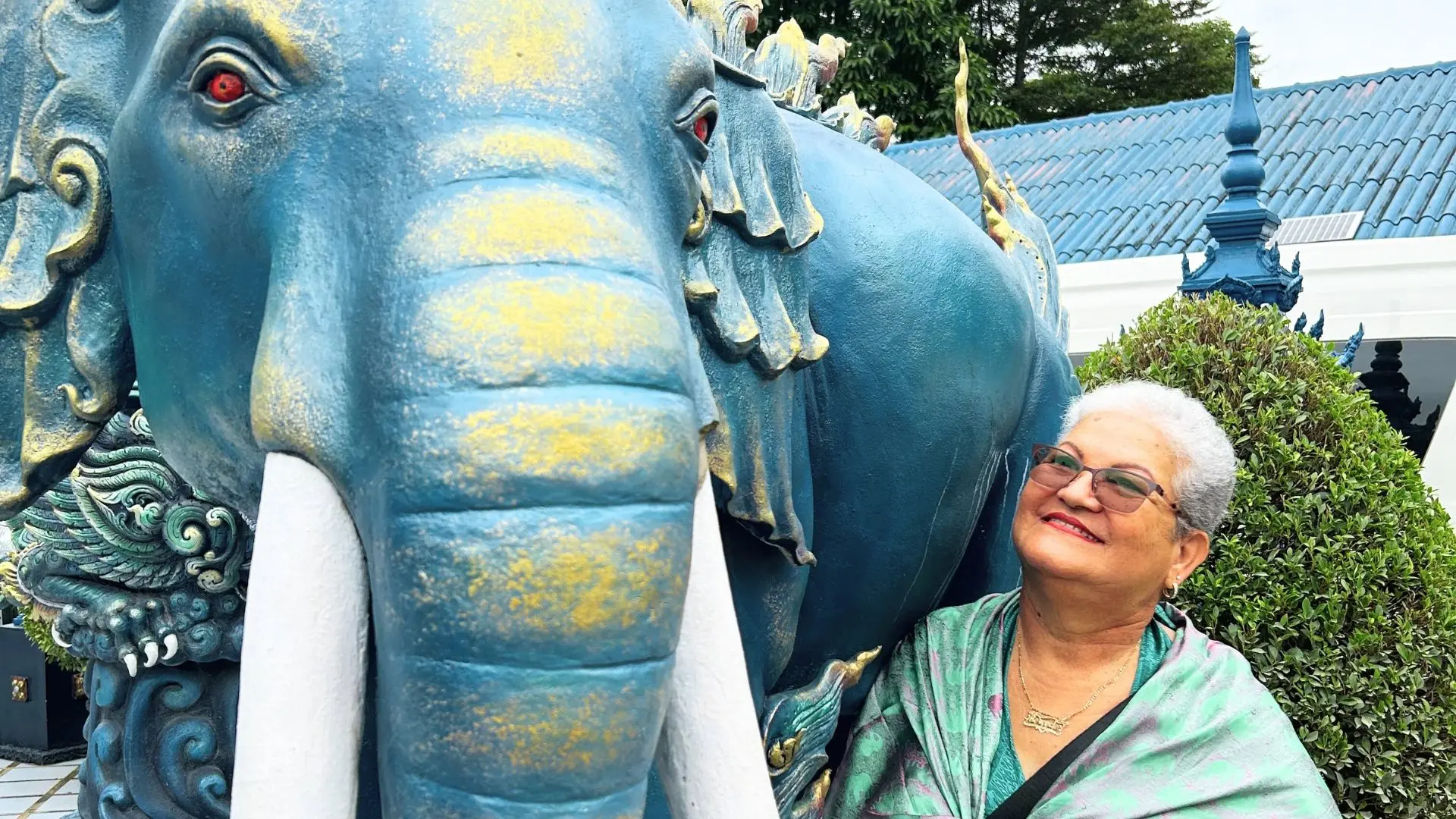 White-haired lady in a green scarf smiles beside the statue of a blue elephant with red eyes