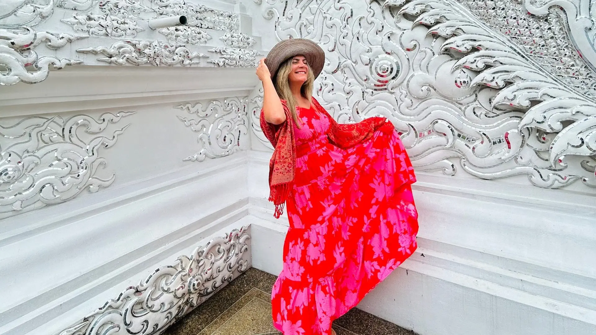 A traveler in a red and pink dress holds her hat before a white wall with reliefs and mirrors