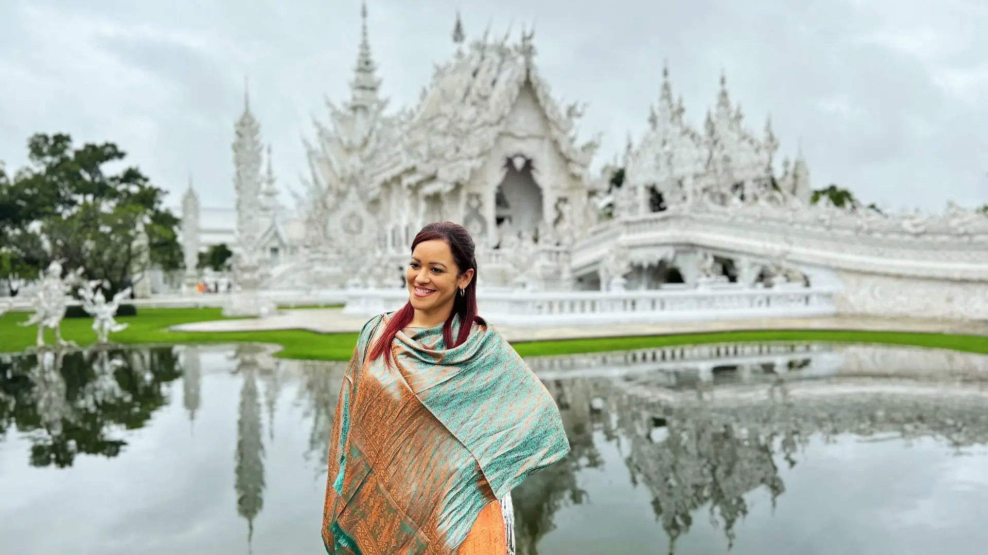 A female traveler in a turquoise and orange shawl smiles by the pond of Wat Rong Khun on a cloudy day