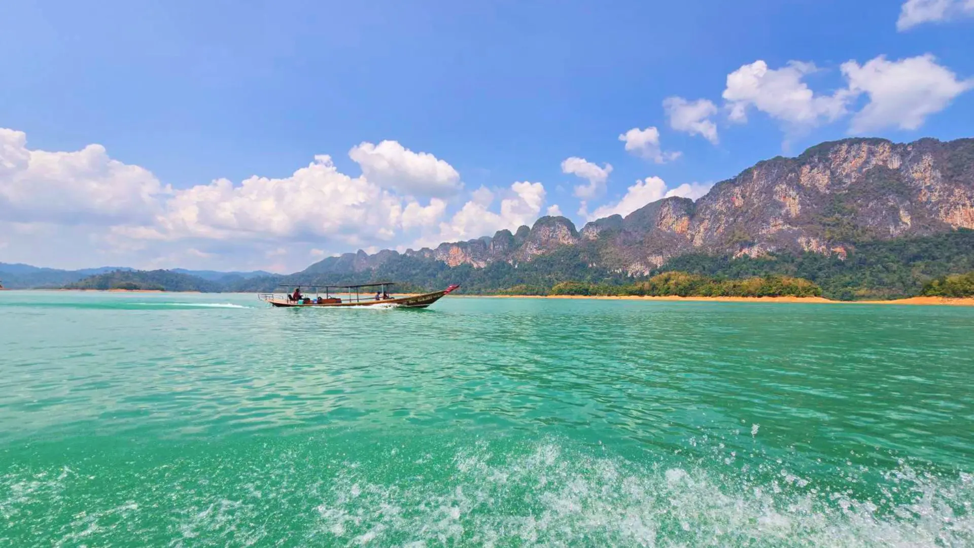 Barco de cola larga navegando por el agua turquesa del lago Cheow Lan frente a montañas de piedra caliza