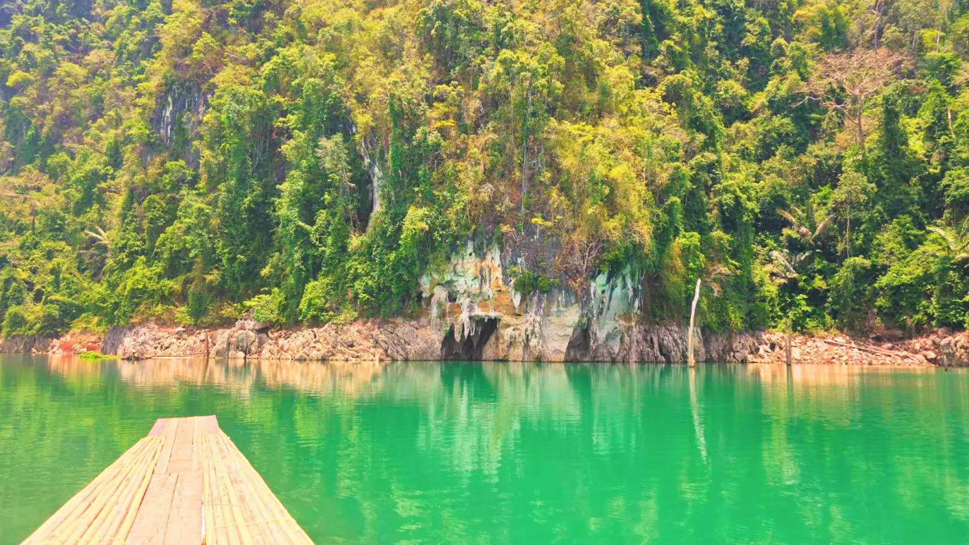 Proa de una balsa de bambú ante un acantilado de selva con la boca de una cueva a ras del agua verde