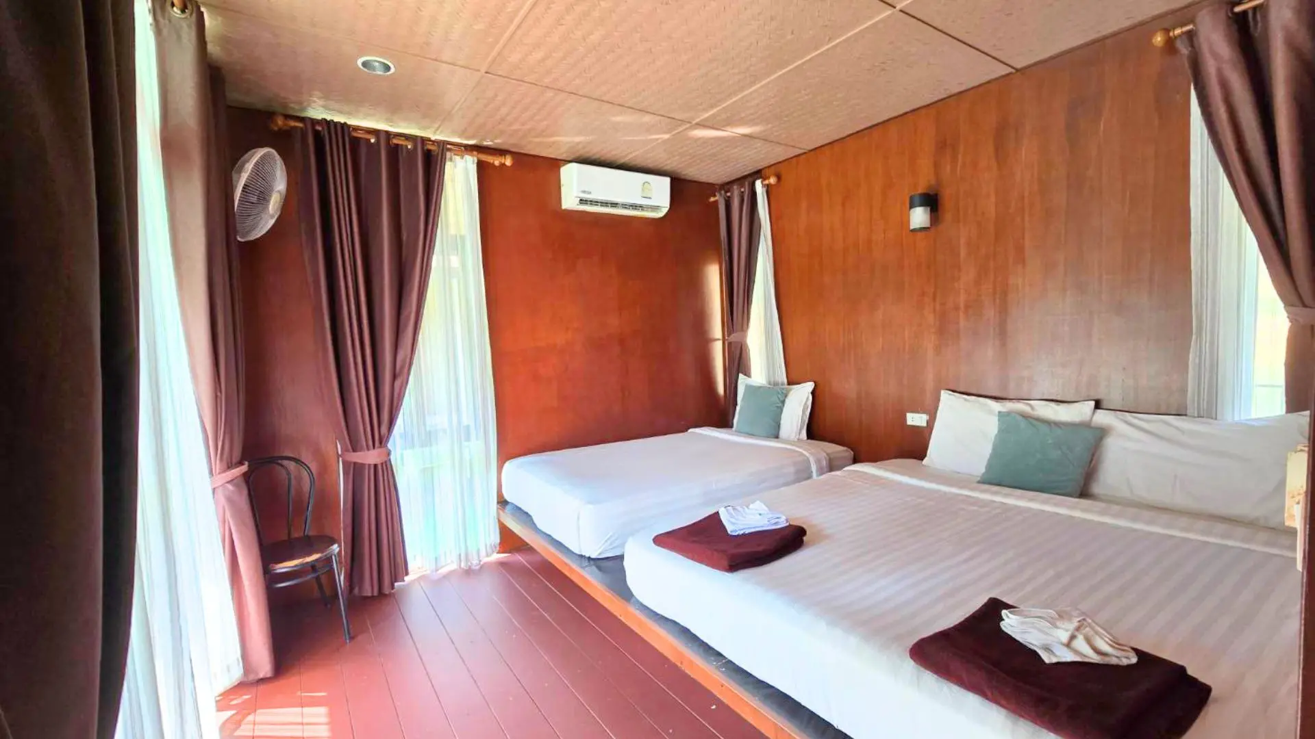Habitación con paredes de madera, dos camas de matrimonio con toallas granates, aire acondicionado y ventilador