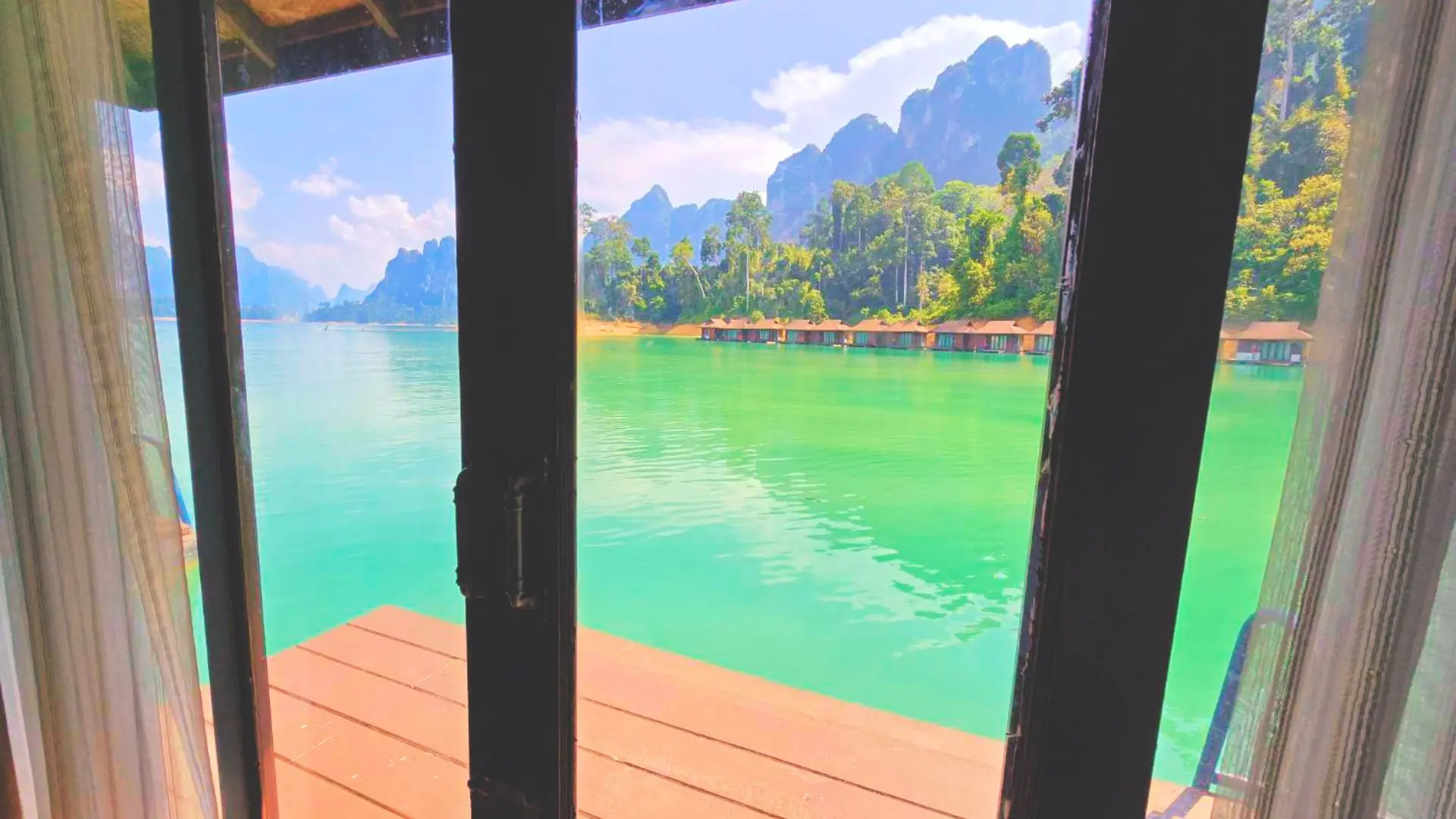Vista desde la puerta de una cabaña flotante: terraza de madera, agua turquesa y más cabañas al pie de montañas calizas