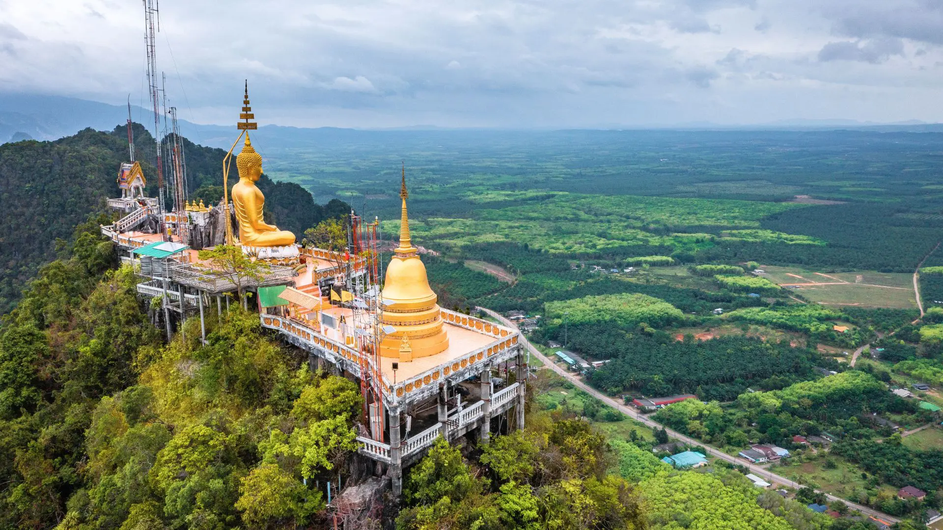 Templo empoleirado num pico de selva com Buda e pagode dourados, e plantações e estrada lá em baixo