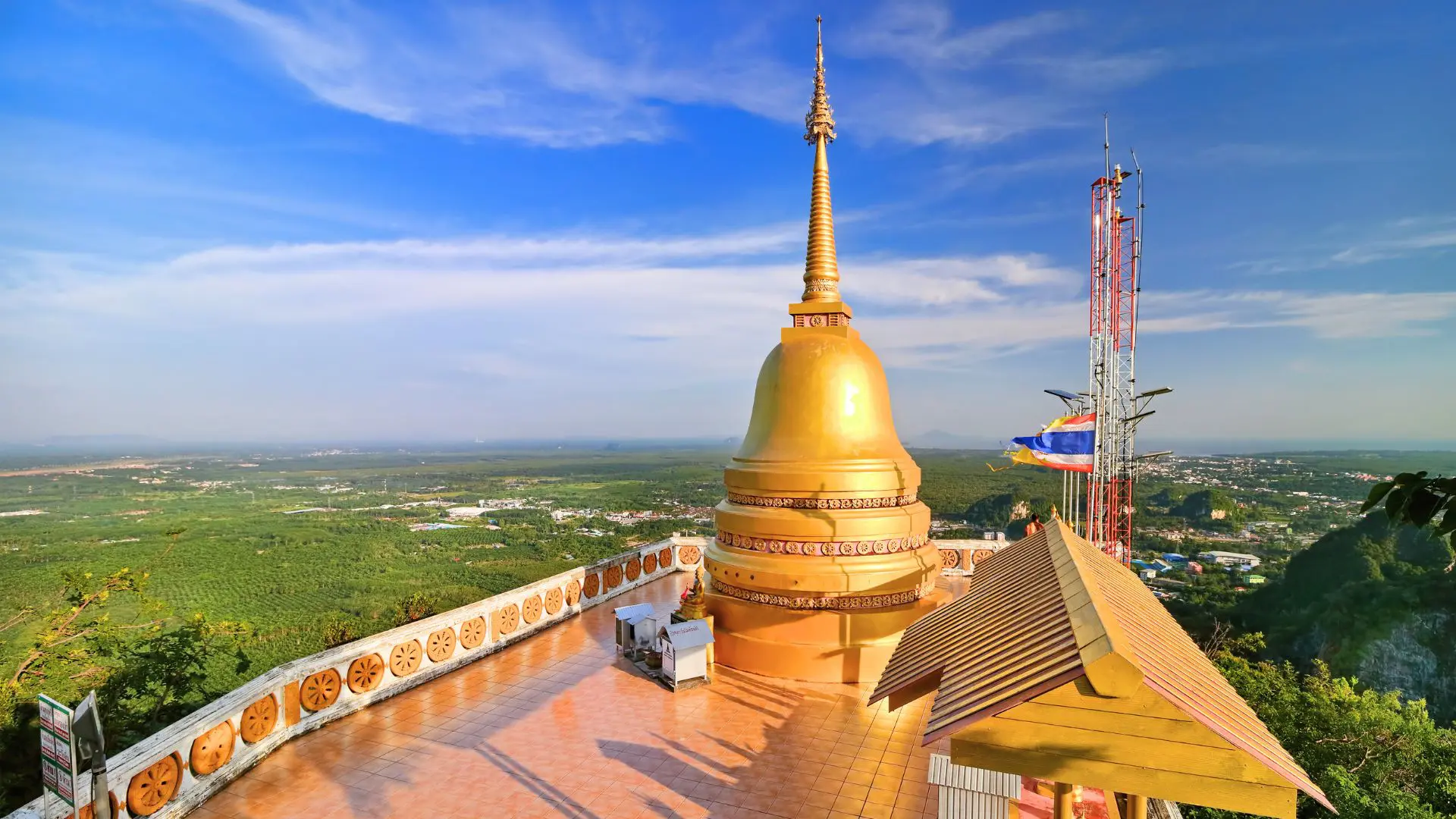 Pagode dourado no terraço do cume, com bandeira da Tailândia, antena e vista para a planície de palmeiras