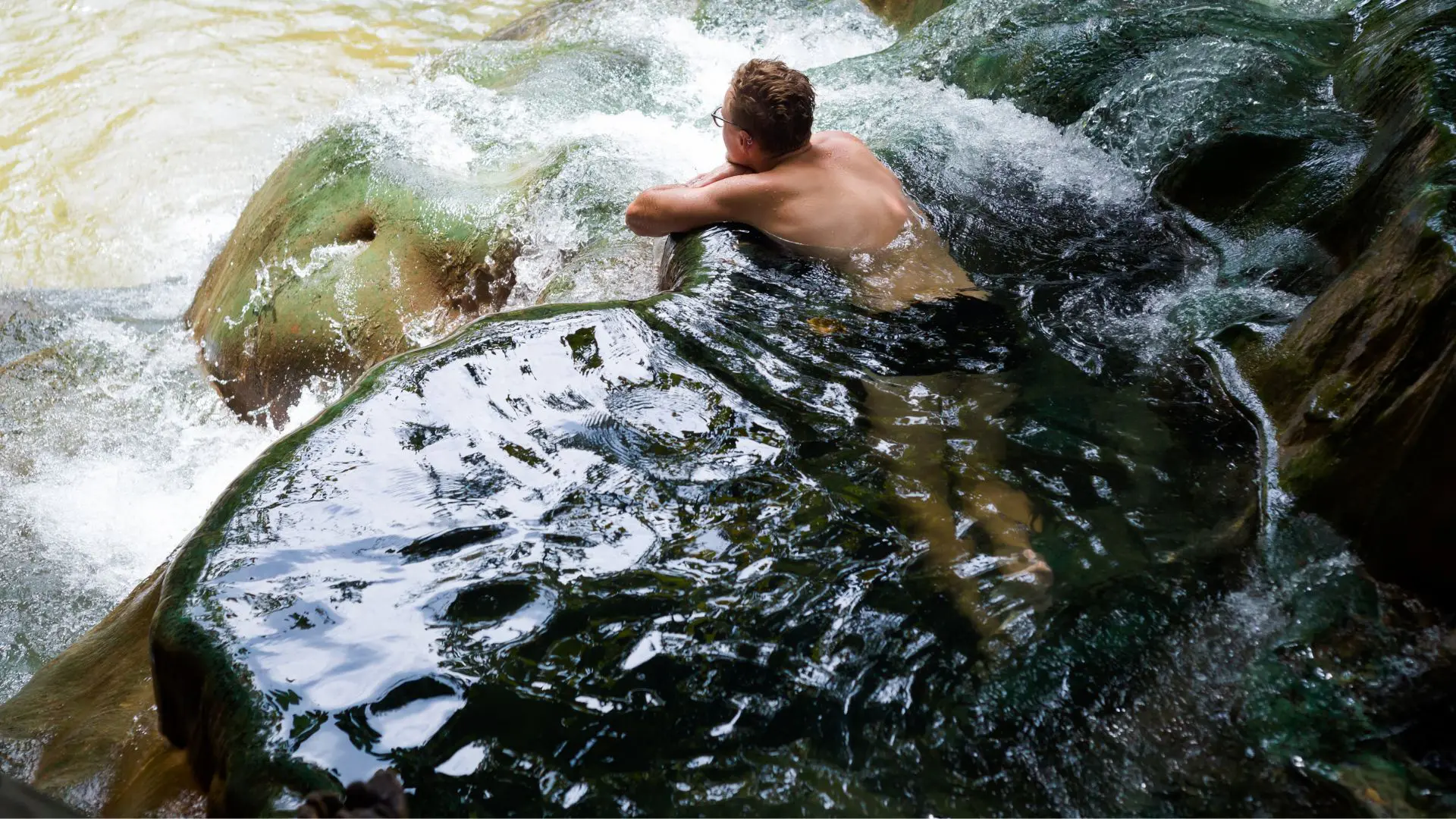 Um homem de óculos banha-se encostado à borda de um poço natural de rocha por onde corre a água