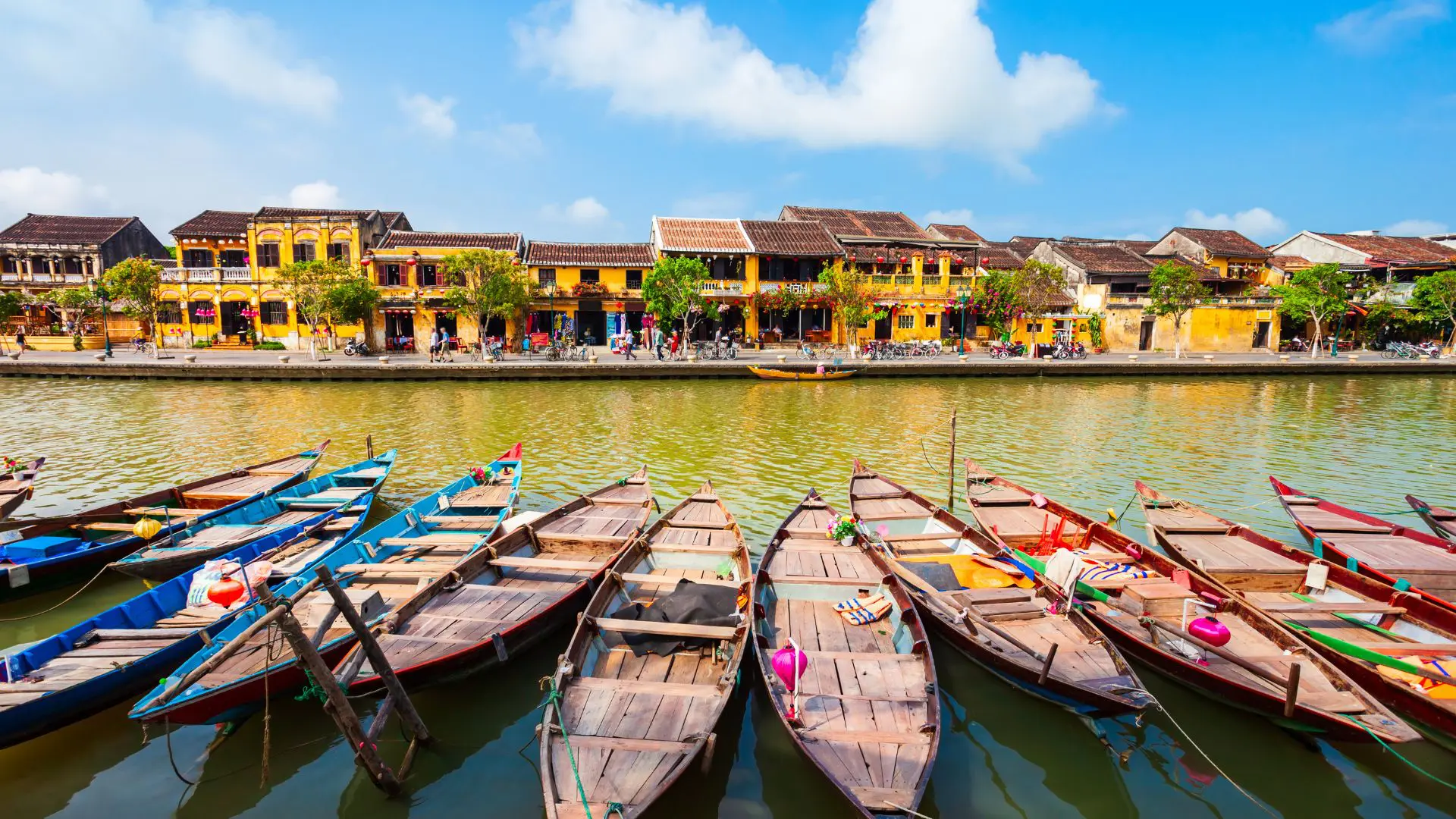 MERCADO DE HOI AN NO VIETNAME (2) Barcos de madeira azuis e vermelhos amarrados em fila diante das casas amarelas de Hoi An, sob um céu azul com nuvens