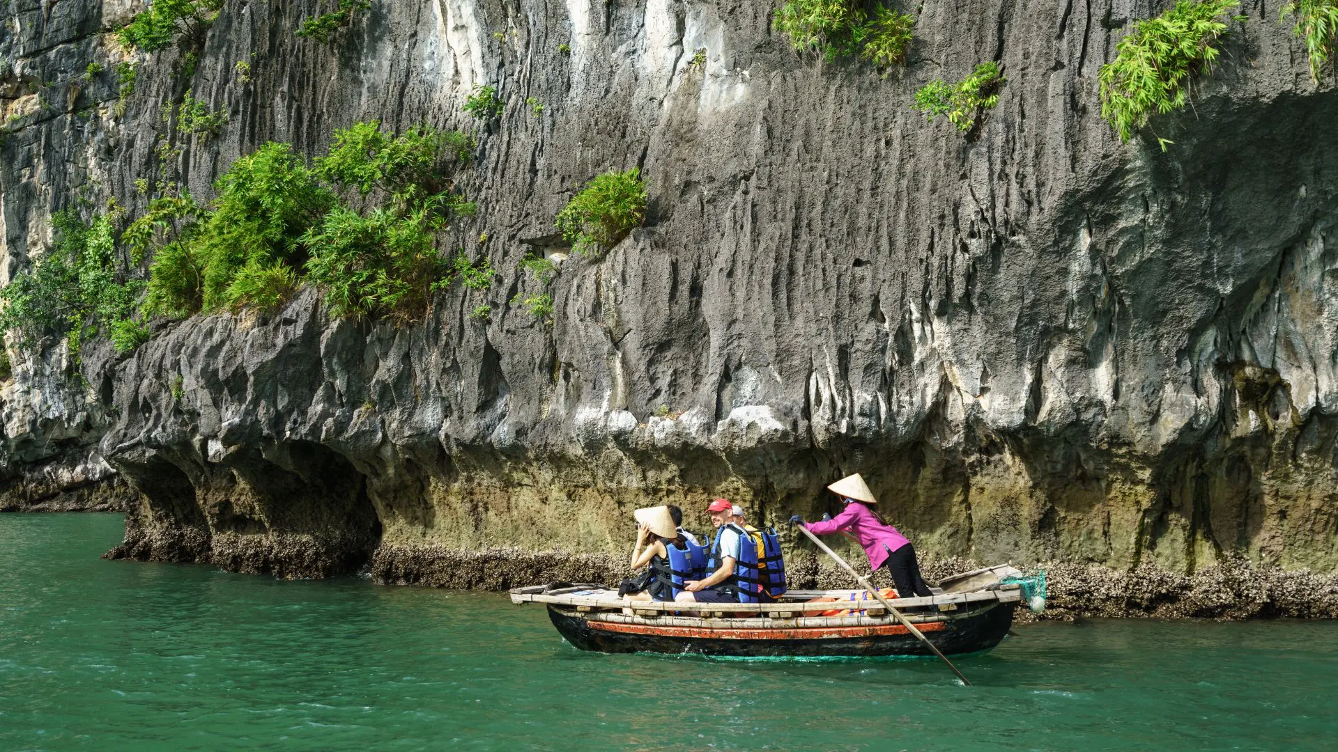 Baía de Halong em Hanói Vietname Barco a remos com três viajantes de colete salva-vidas e uma remadora de chapéu cónico, junto a uma falésia rochosa