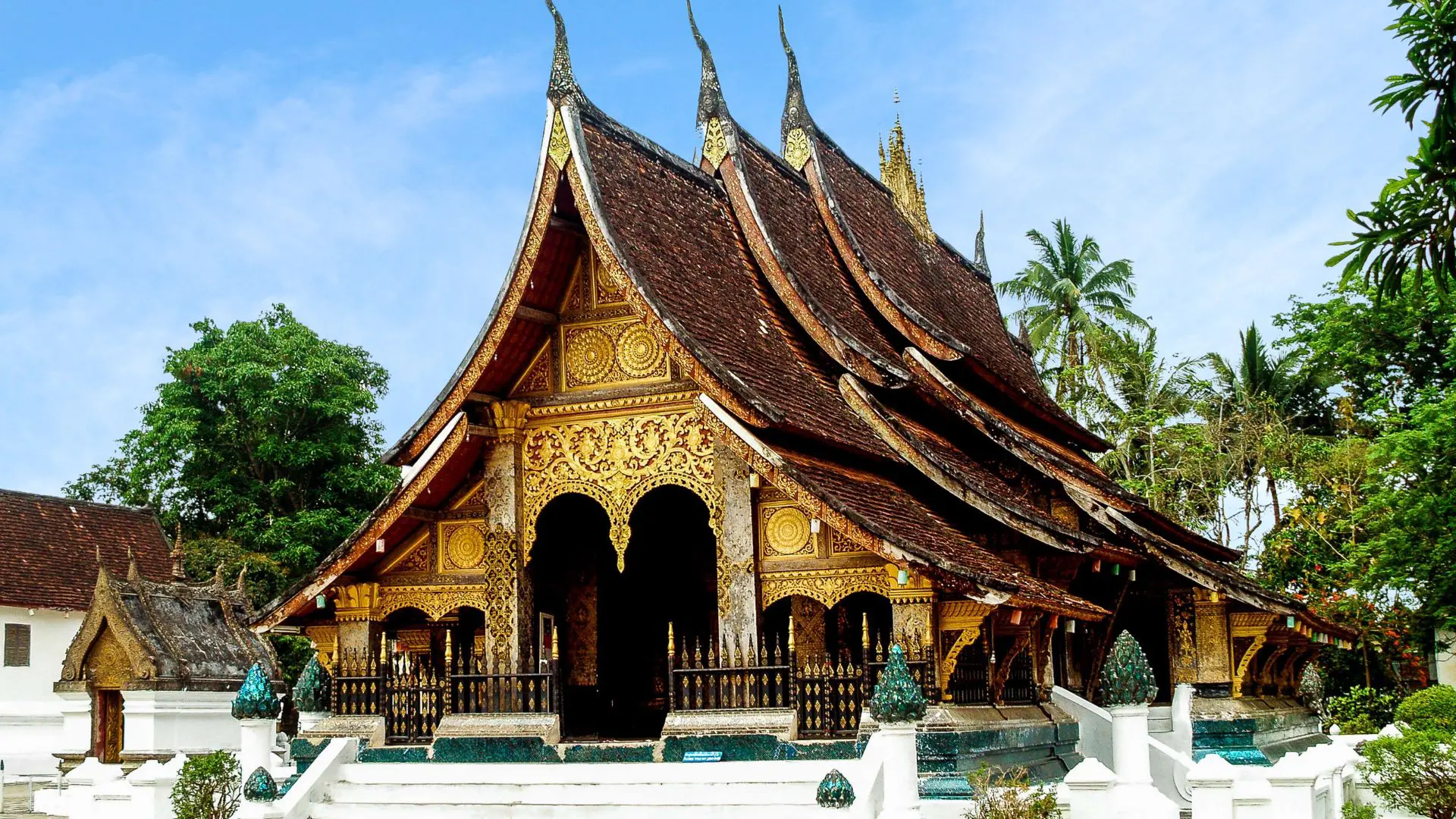Templo laosiano de telhados curvos em cascata e fachada dourada, o Wat Xieng Thong, rodeado de palmeiras