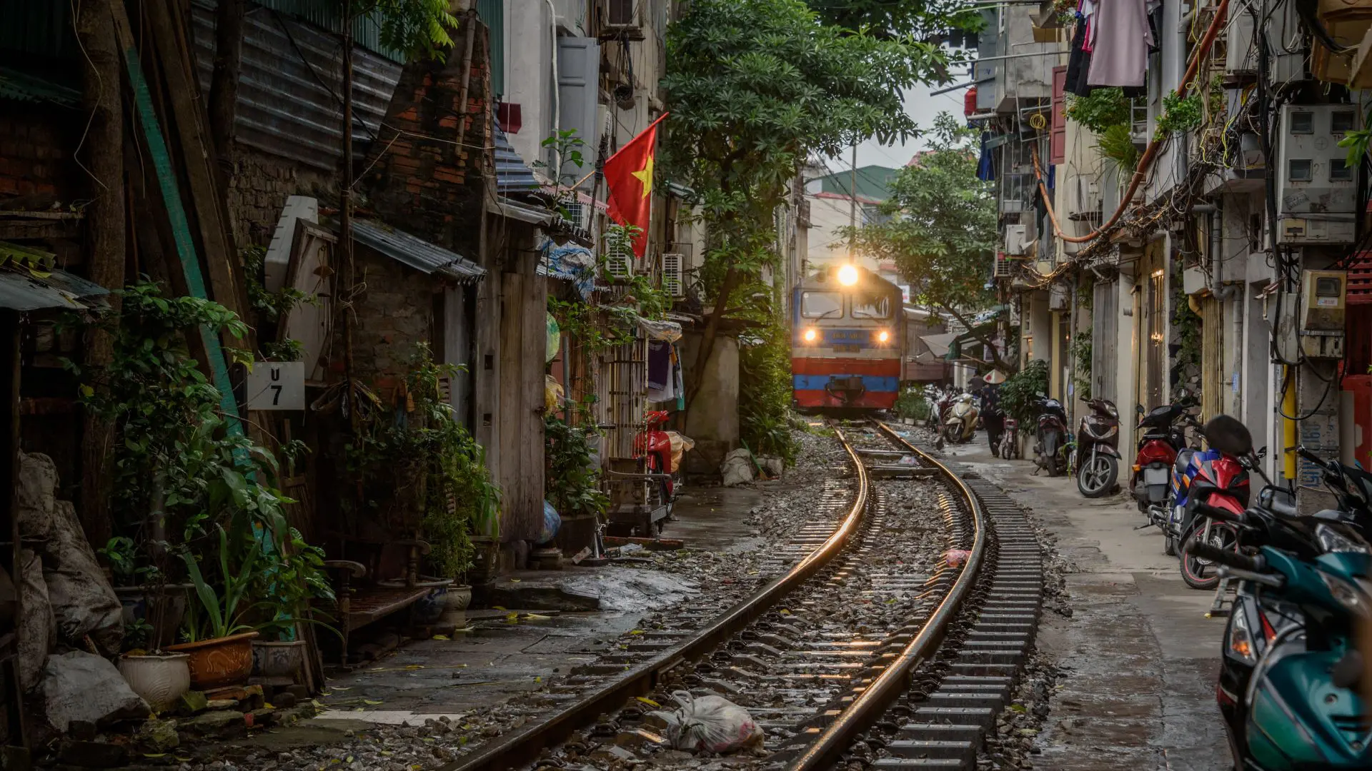 Comboio em Hanói Vietname Comboio de farol aceso avança por uma rua estreita entre casas, com motas estacionadas e uma bandeira do Vietname