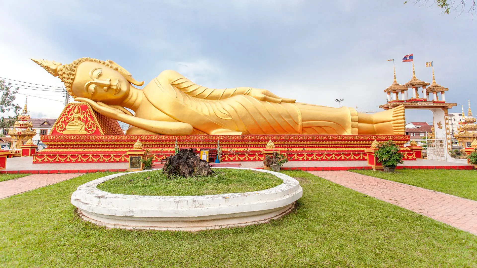 Buda reclinado dourado sobre uma base vermelha e dourada, com relva à frente e a bandeira do Laos a ondular ao fundo