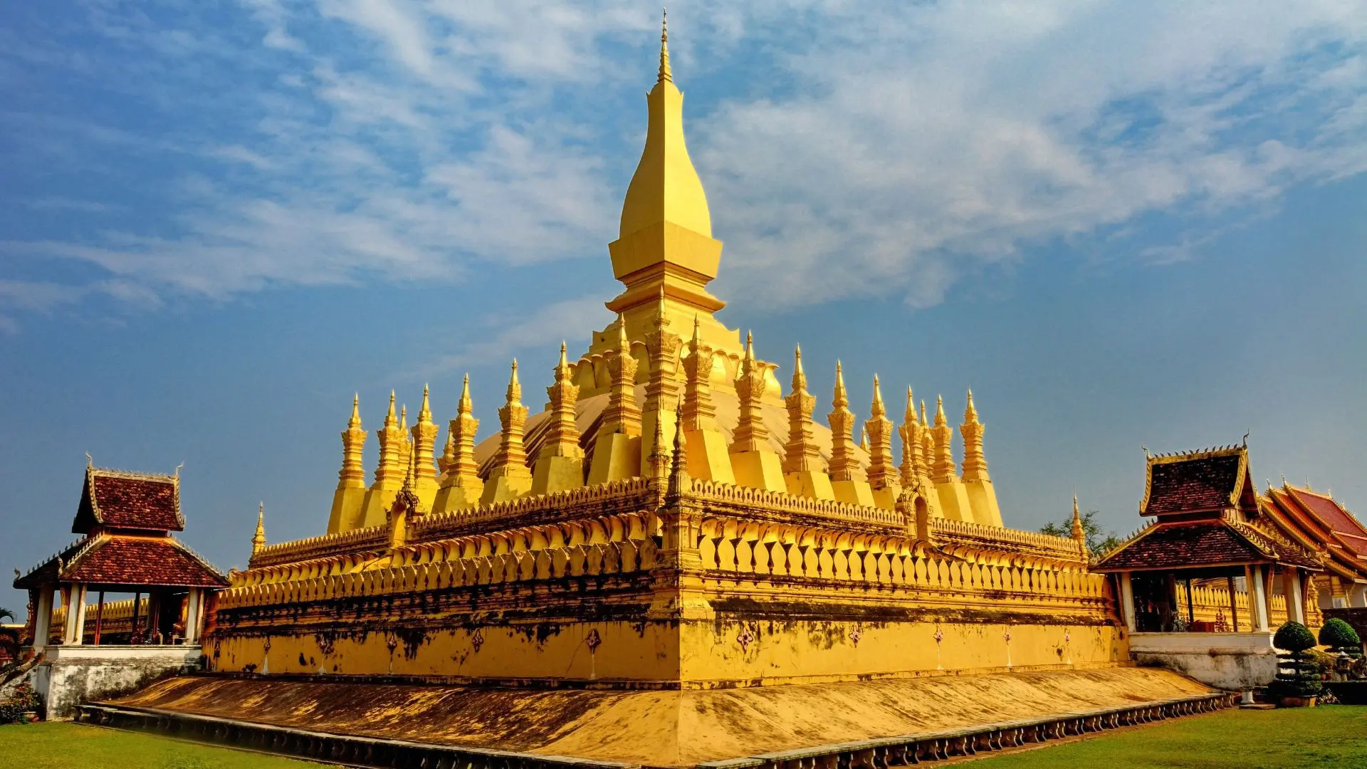 Grande estupa dourada de Pha That Luang com os seus pináculos, entre dois pavilhões de telhado vermelho, em Vientiane