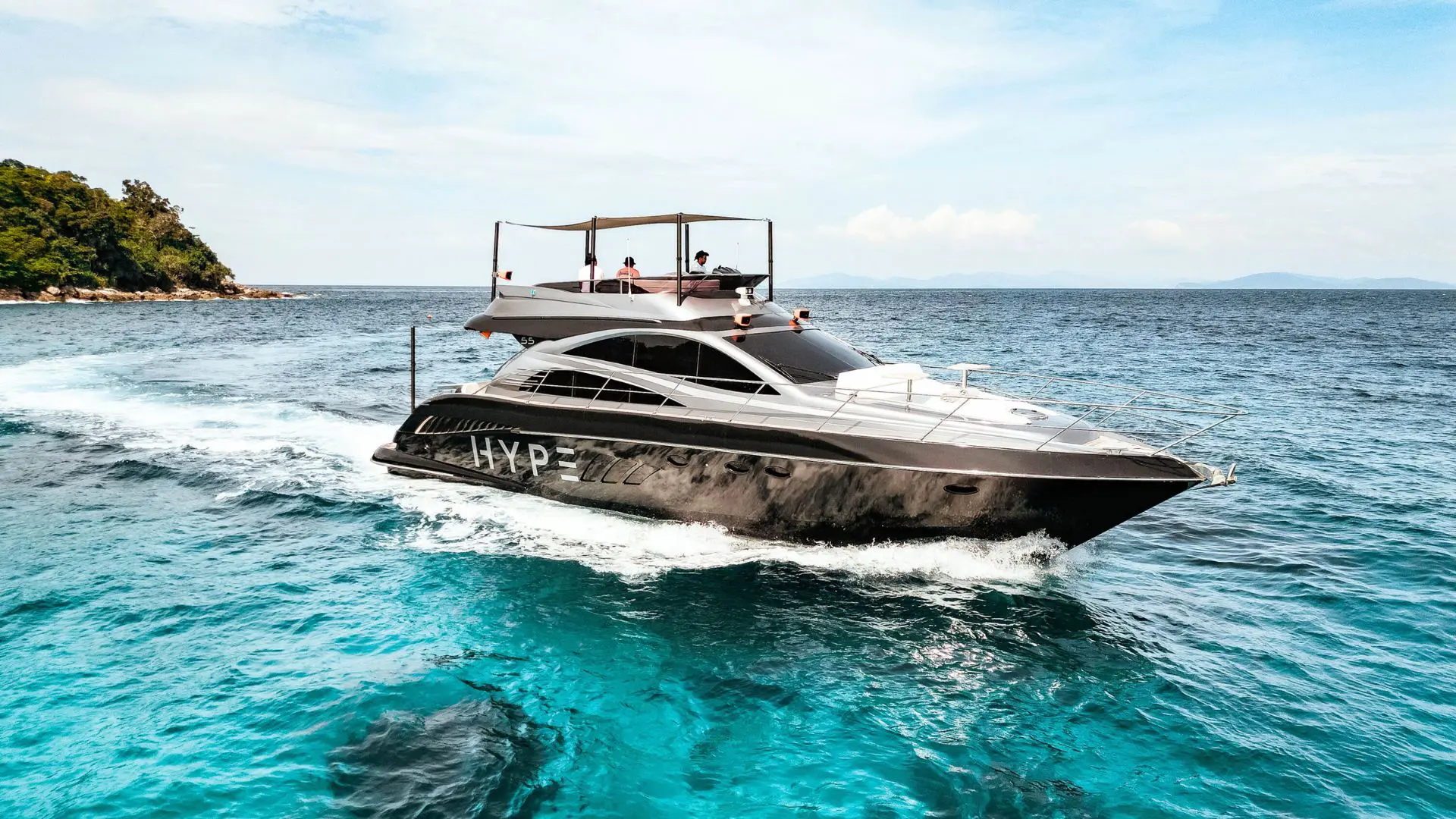 Super Yacht en Phuket (2) Black yacht with the HYPE lettering sailing on turquoise water, three passengers on the upper deck under the canopy