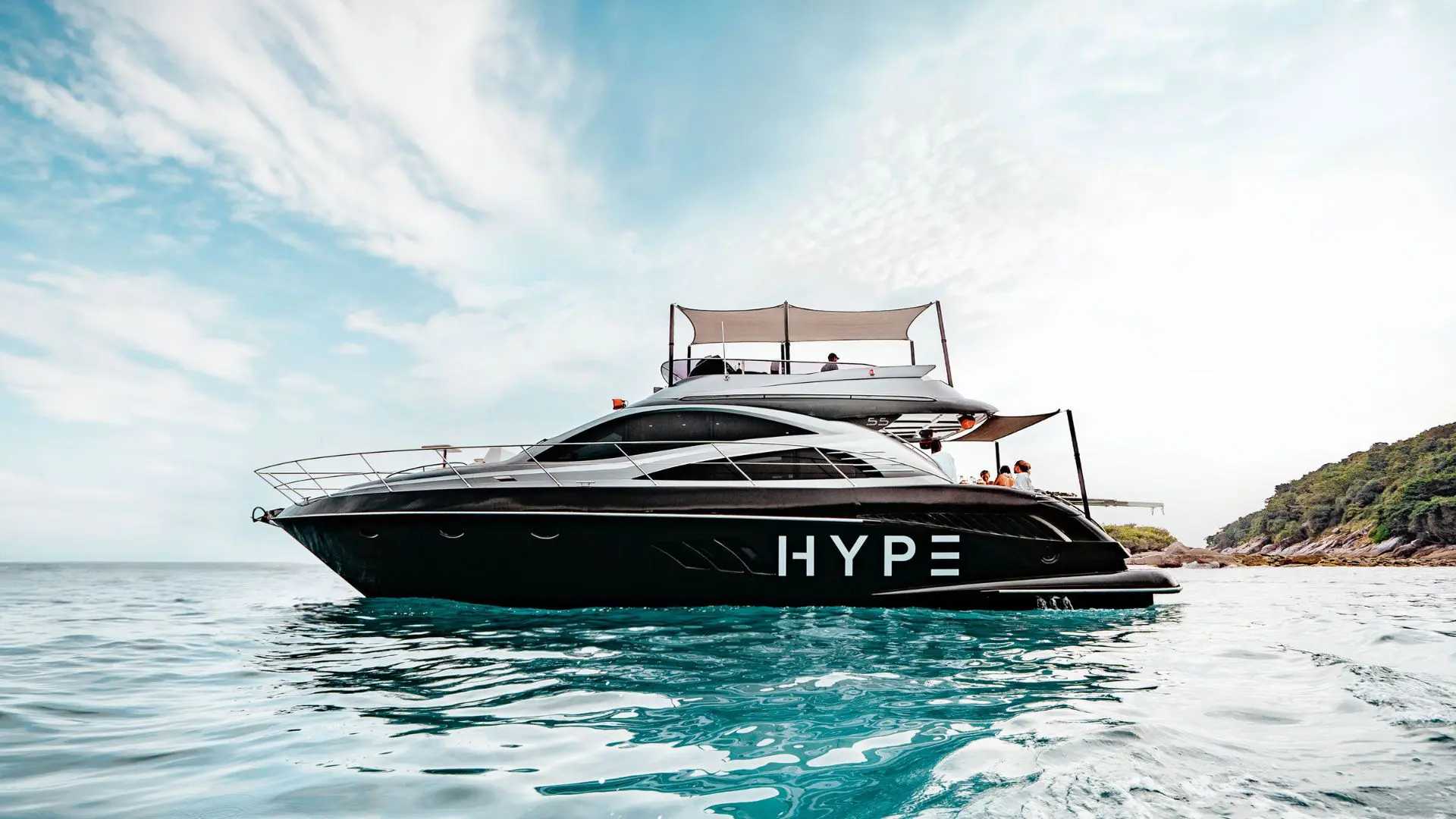 Yate negro con letras HYPE fondeado junto a una costa boscosa, con pasajeros en la popa bajo los toldos