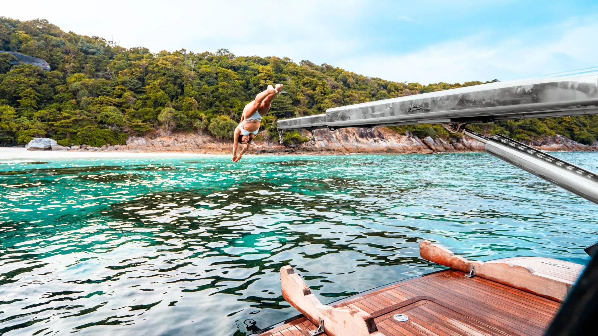 Mujer en bikini salta de cabeza al agua turquesa desde un yate con cubierta de teca, frente a una costa boscosa