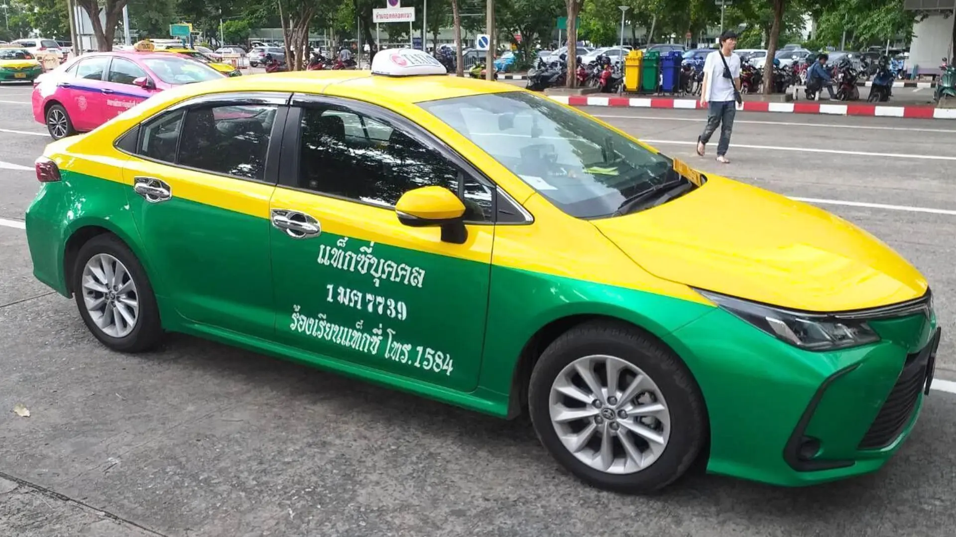 Táxi tailandês verde e amarelo estacionado numa rua com árvores e motas ao fundo